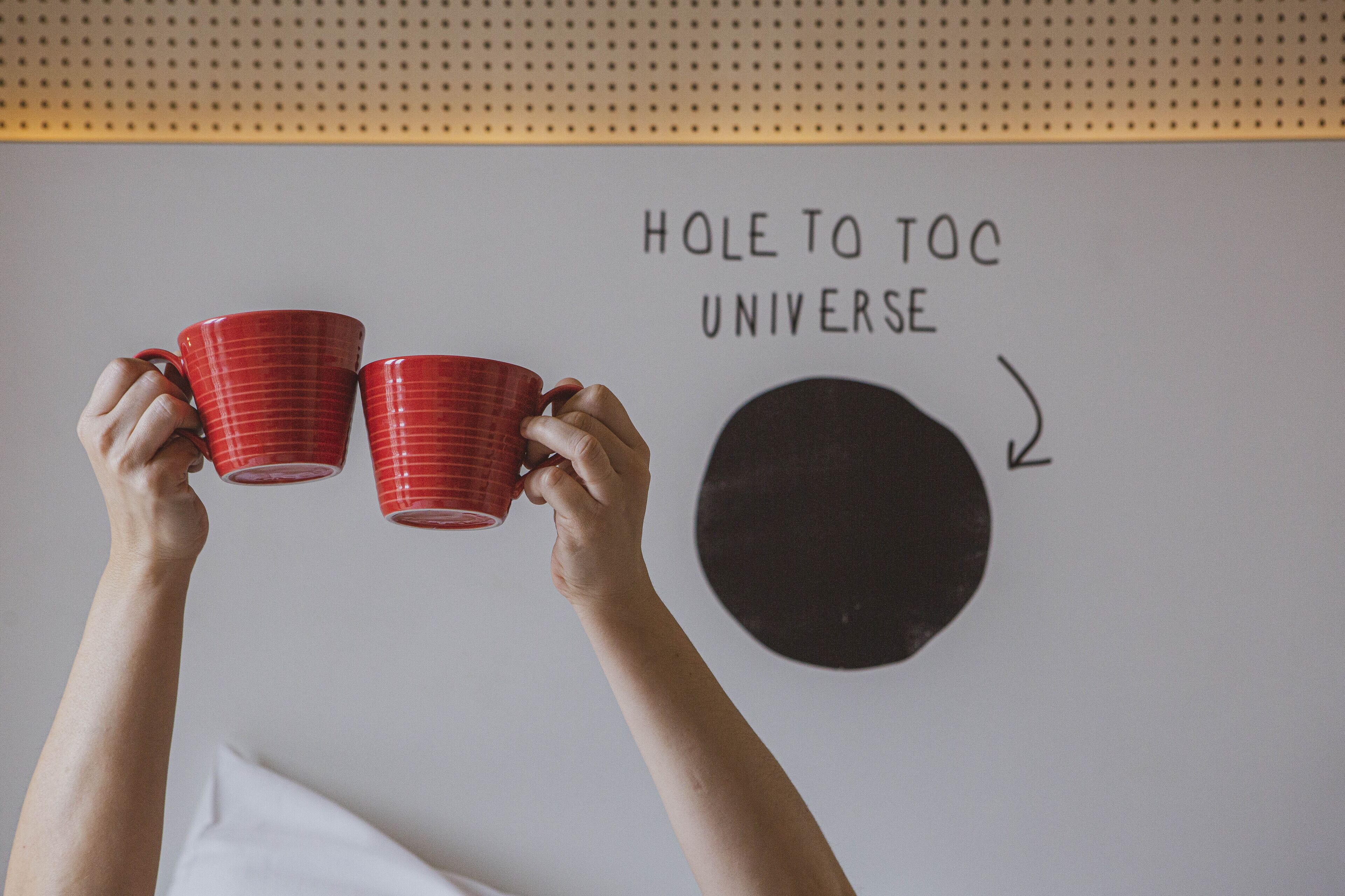 A person holding two red mugs with a wall that has a circular design and the text "HOLE TO TOC UNIVERSE".