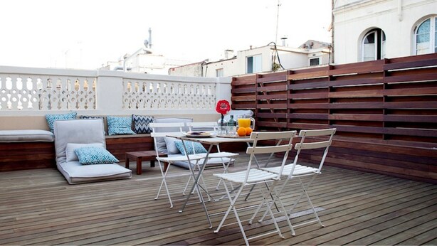 A rooftop terrace with a wooden deck, seating area with cushions, a table with chairs, and a decorative fence. - TOC Hostel Barcelona (Barcelona)