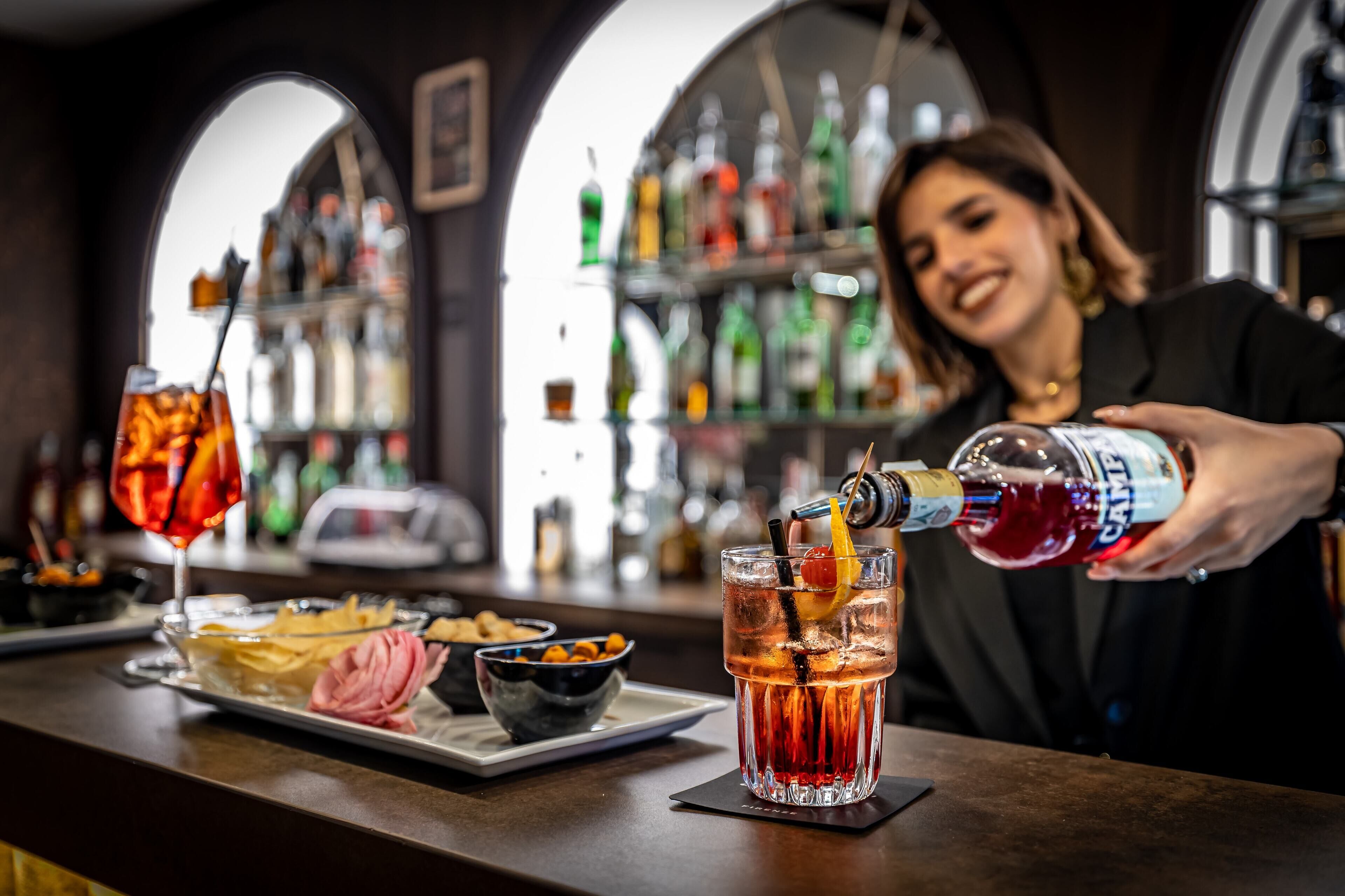 A bar setting with a bartender pouring a drink into a glass, a plate with snacks, and a variety of bottles in the background.