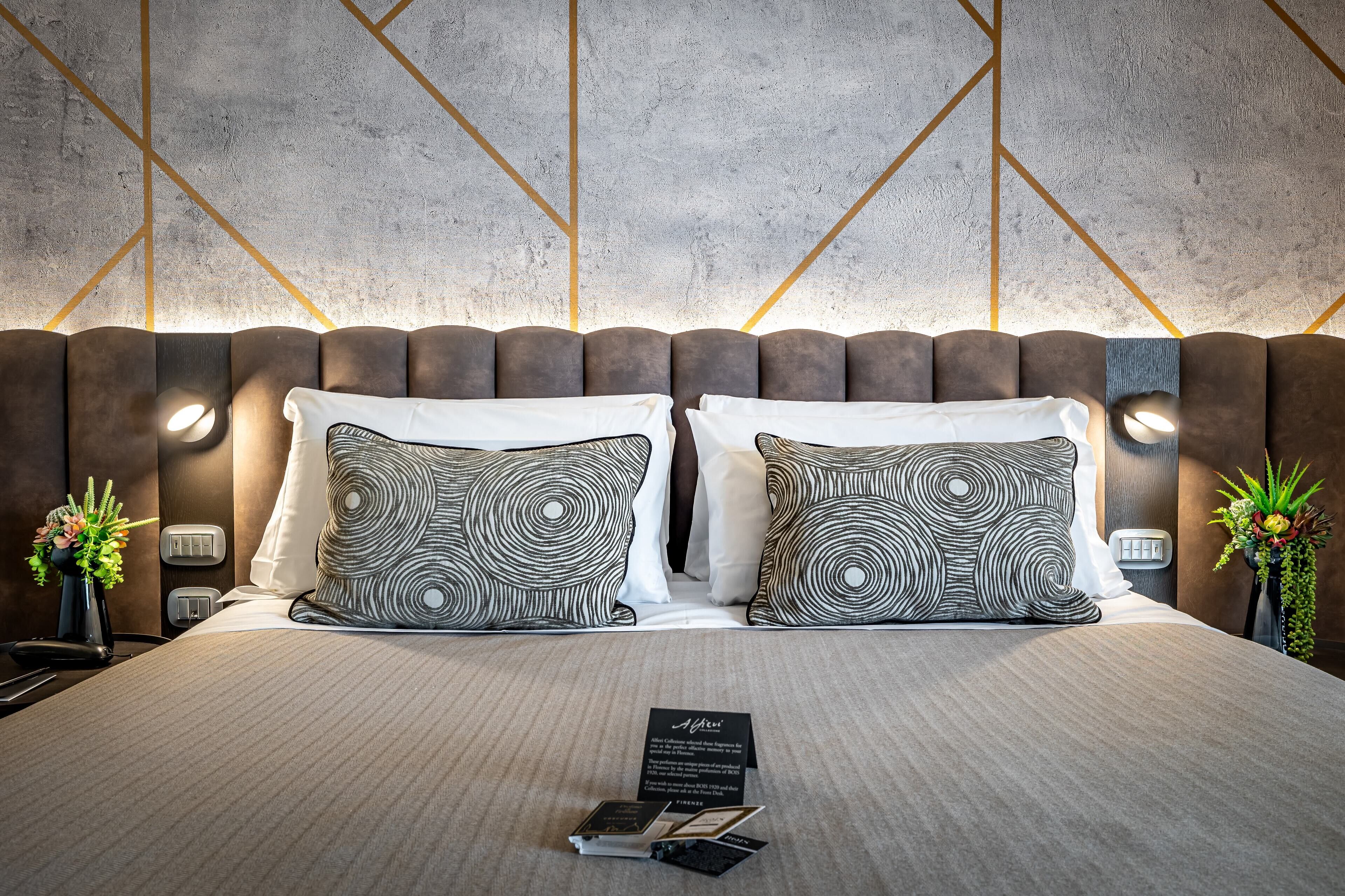 A modern hotel room with a large bed, two patterned pillows, a bedside table with a lamp, and a wall with geometric patterns.