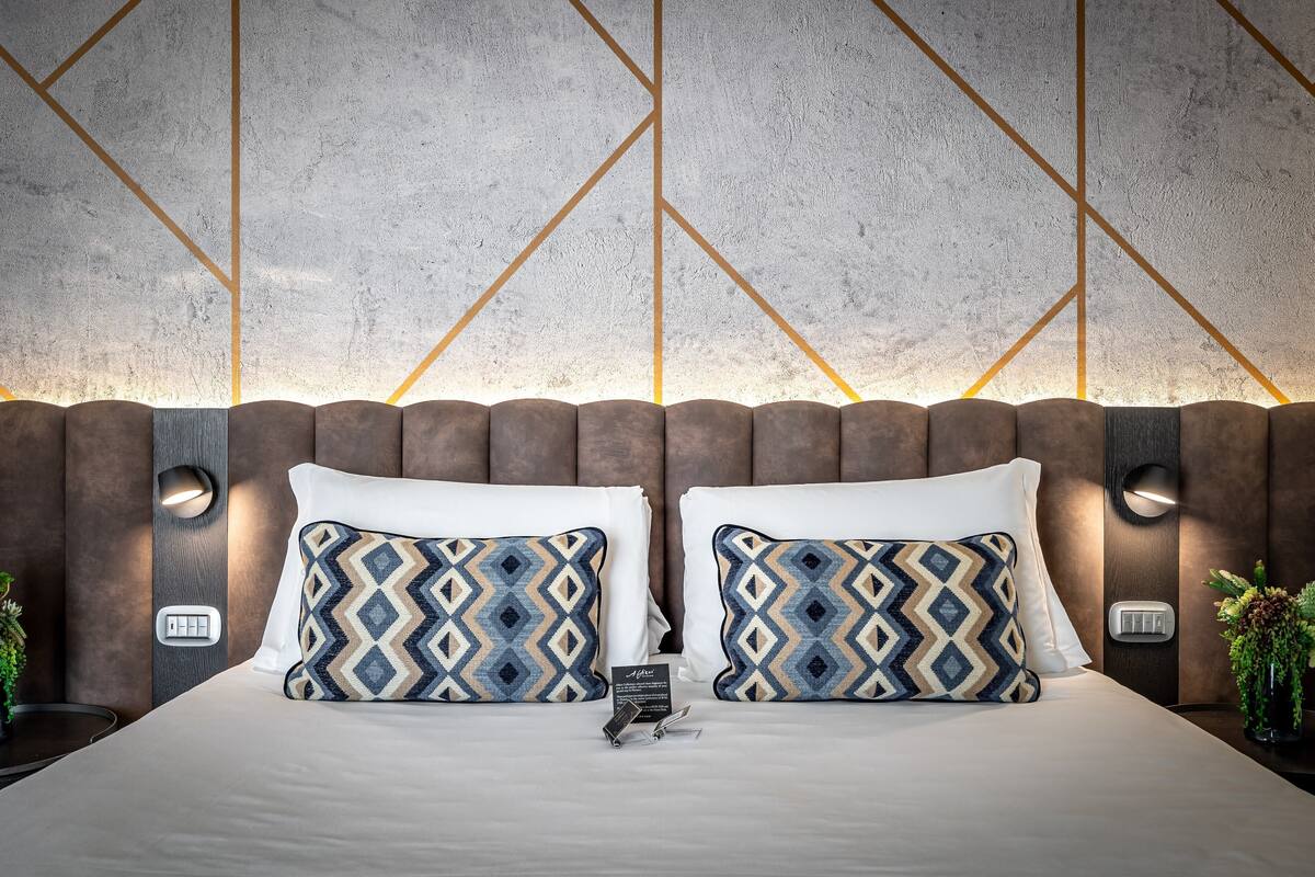 Camera d'albergo moderna con un ampio letto, due cuscini coordinati a motivi decorativi, una testiera in legno e illuminazione a parete.