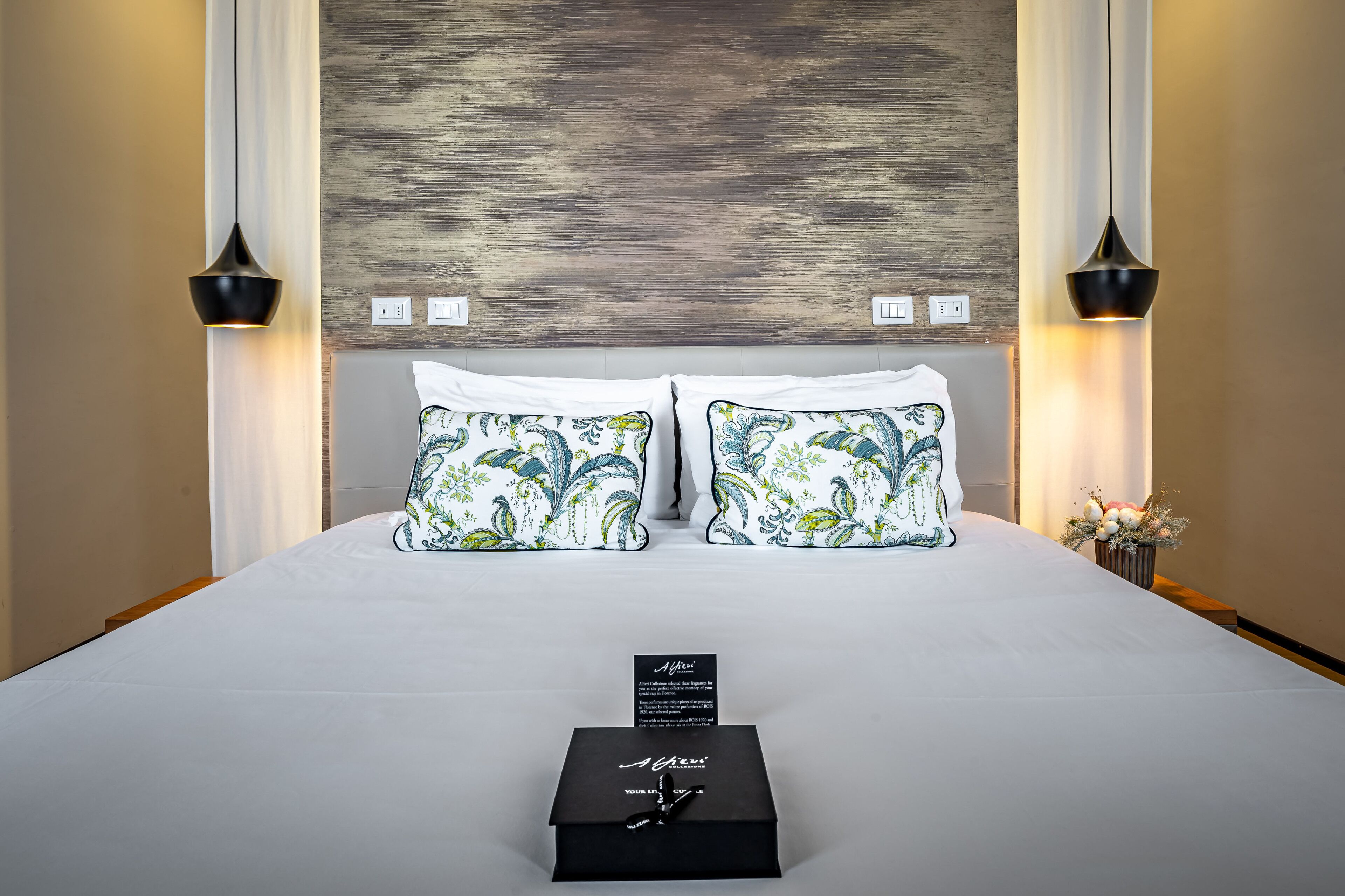 Una cama bien tendida con almohadas estampadas, una caja negra sobre la cama y dos lámparas fijadas a la pared.