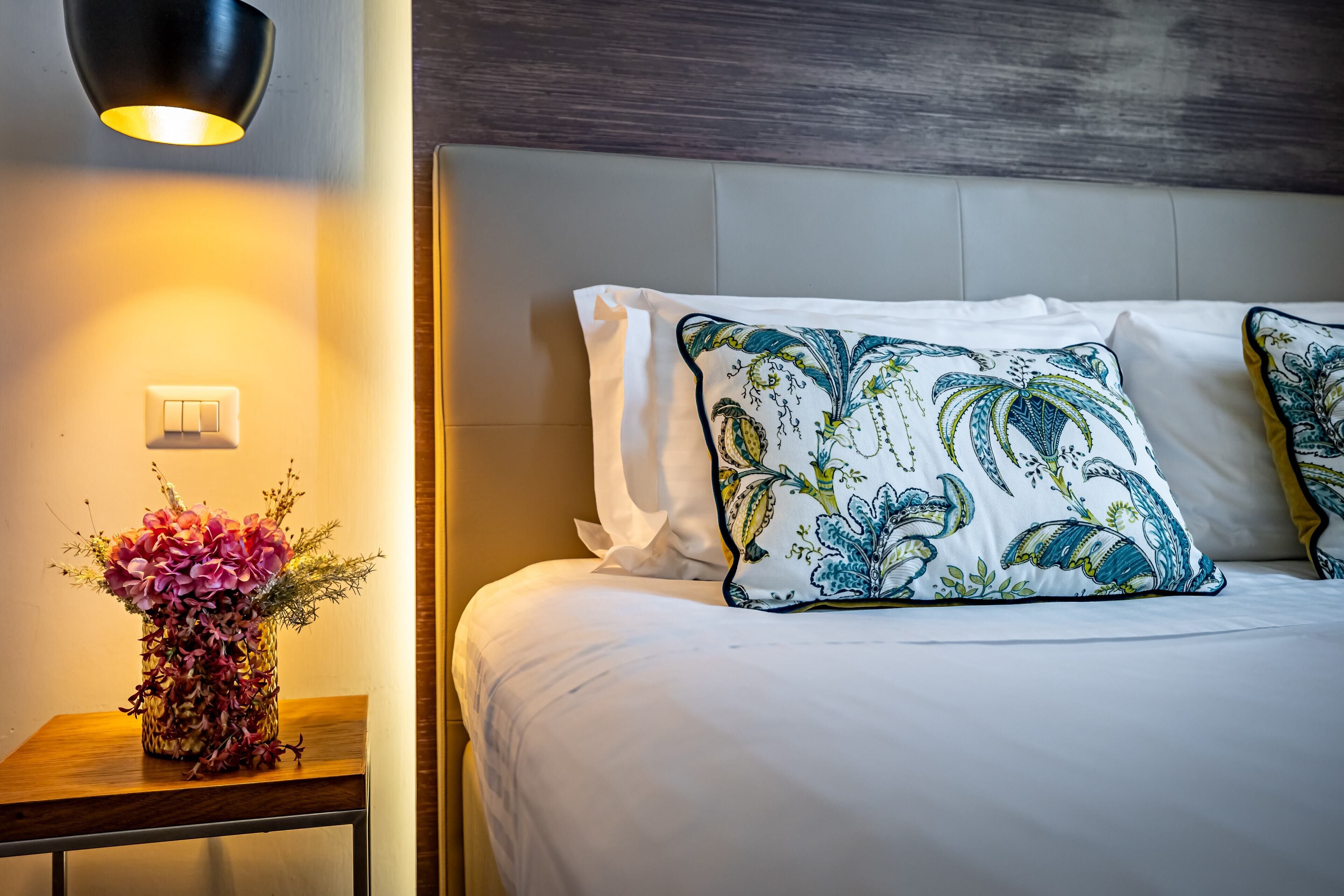 A neatly made bed with white linens and decorative pillows, a bedside table with a vase of flowers, and a wall-mounted light fixture.