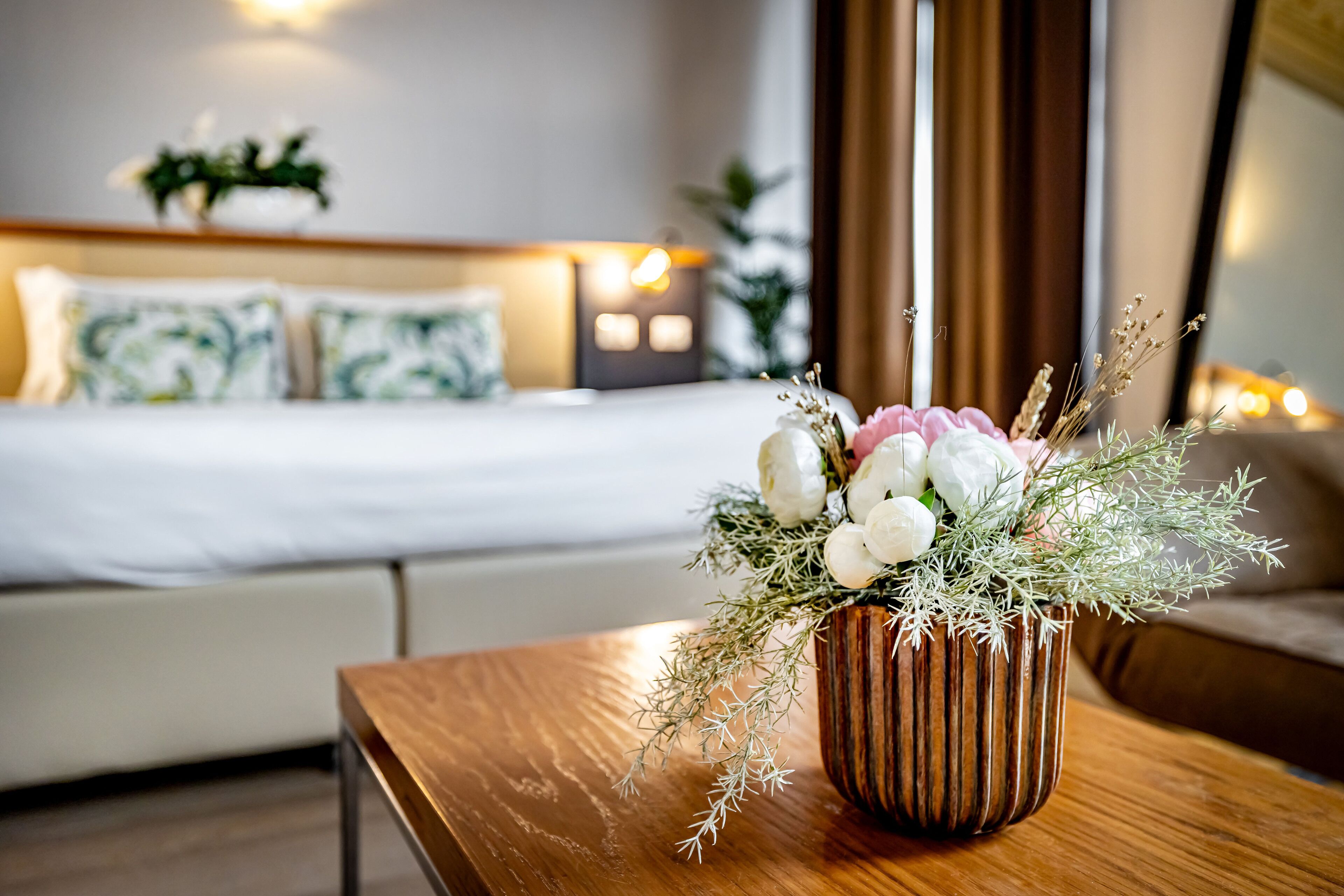 A hotel room with a neatly made bed, a wooden side table with a floral arrangement, and a nightstand with a lamp.