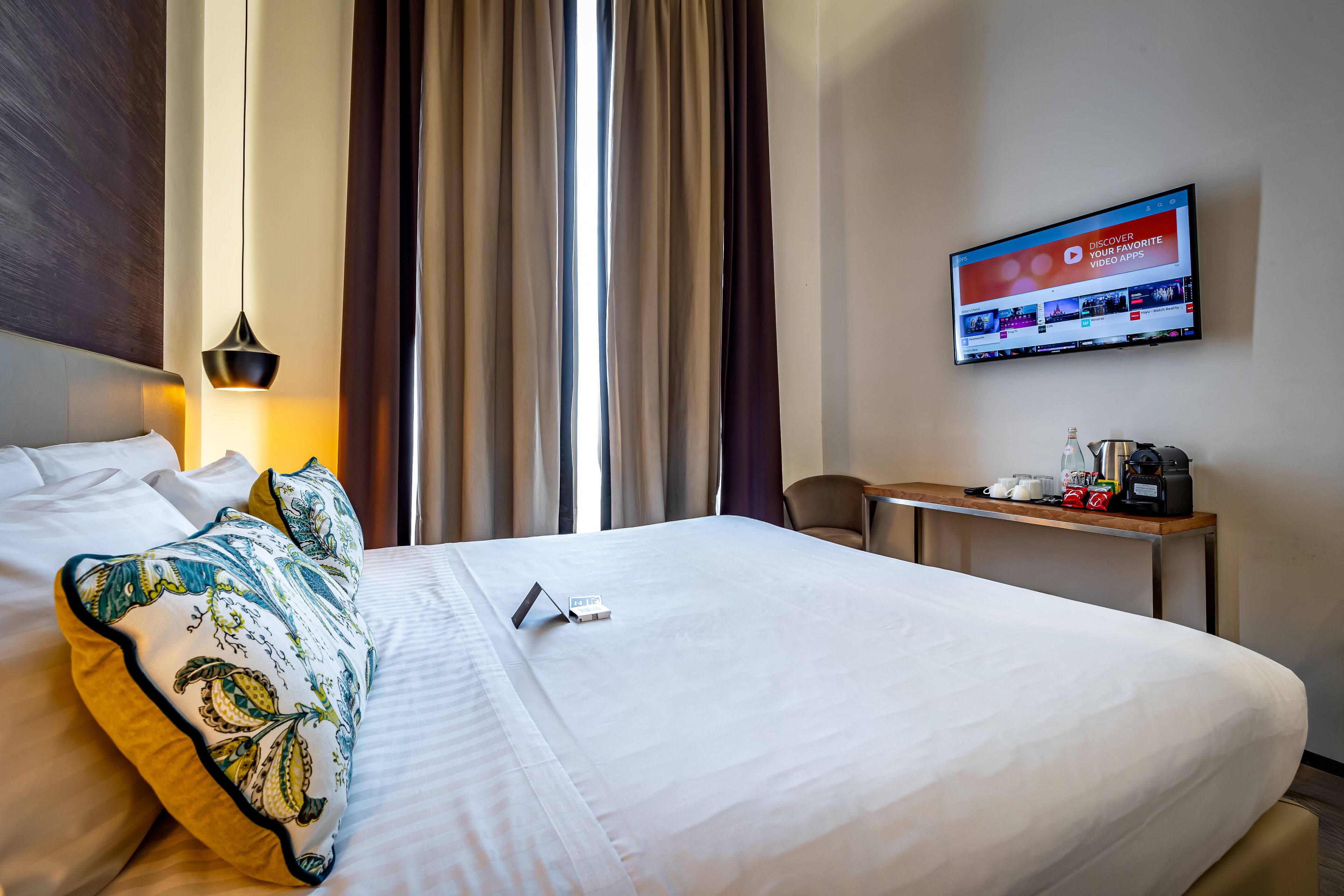 Une chambre d’hôtel avec un grand lit, une table de chevet avec une machine à café, un téléviseur affichant une application vidéo et une fenêtre avec des rideaux.