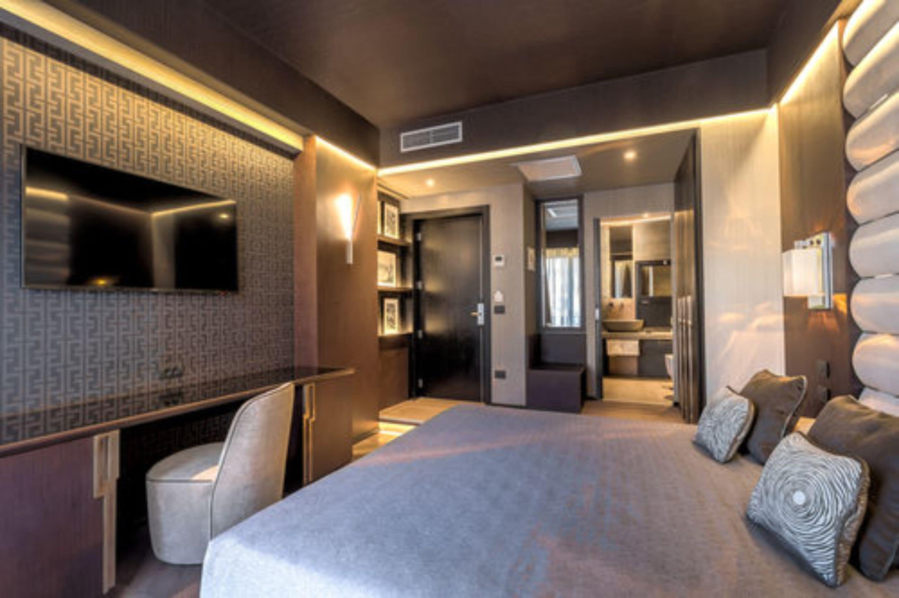 Ein modernes Hotelzimmer mit einem großen Bett, einem Schreibtisch, einem Flachbildfernseher und einem durchgehenden Türspalt einsehbarer Badezimmer.