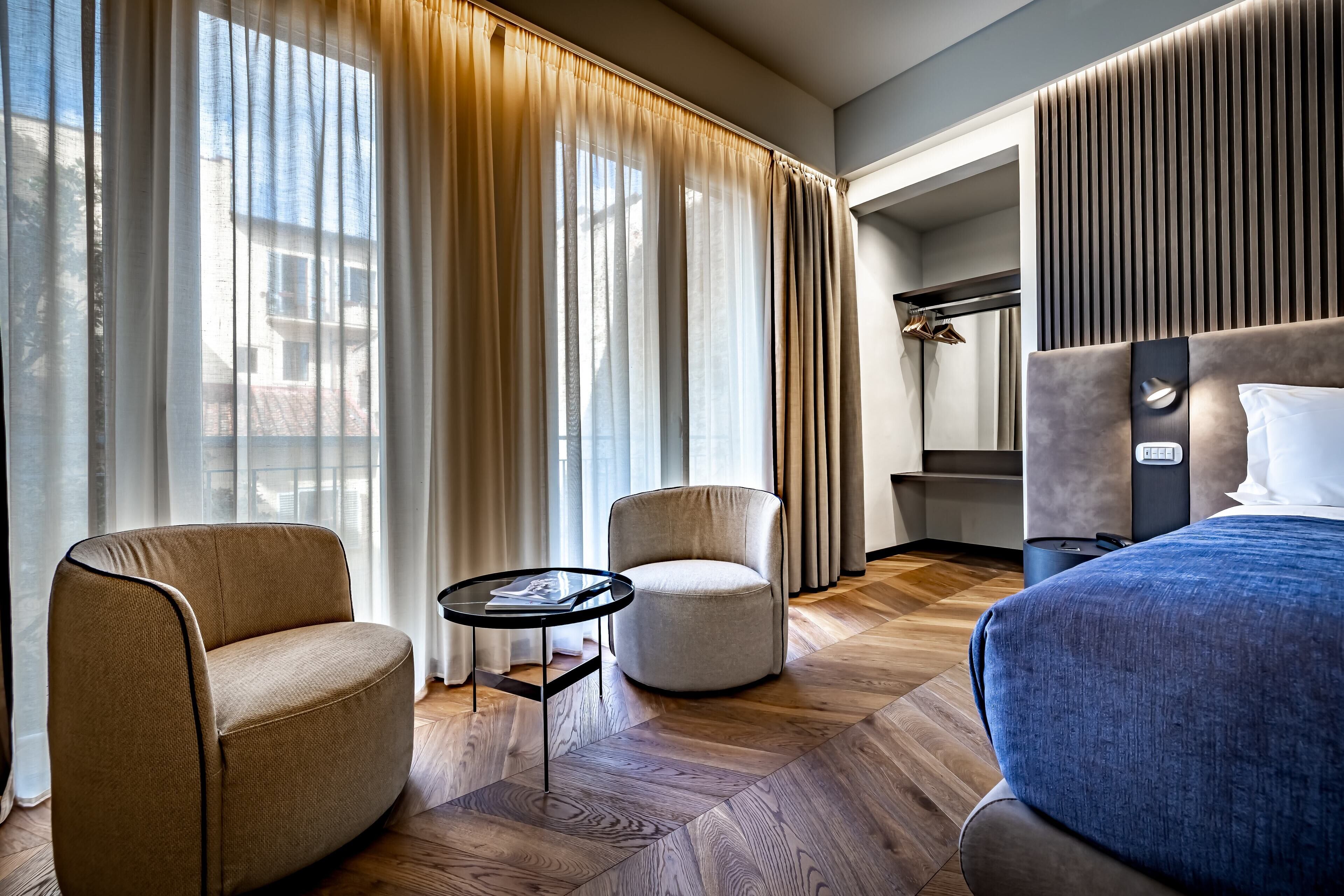 Ein modernes Hotelzimmer mit einem großen Bett, zwei Sesseln, einem kleinen Tisch und Blick durch leichte Vorhänge.