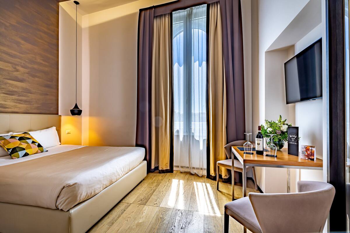 Una camera d'albergo con un letto, una scrivania, una sedia, una TV e una finestra con tende.