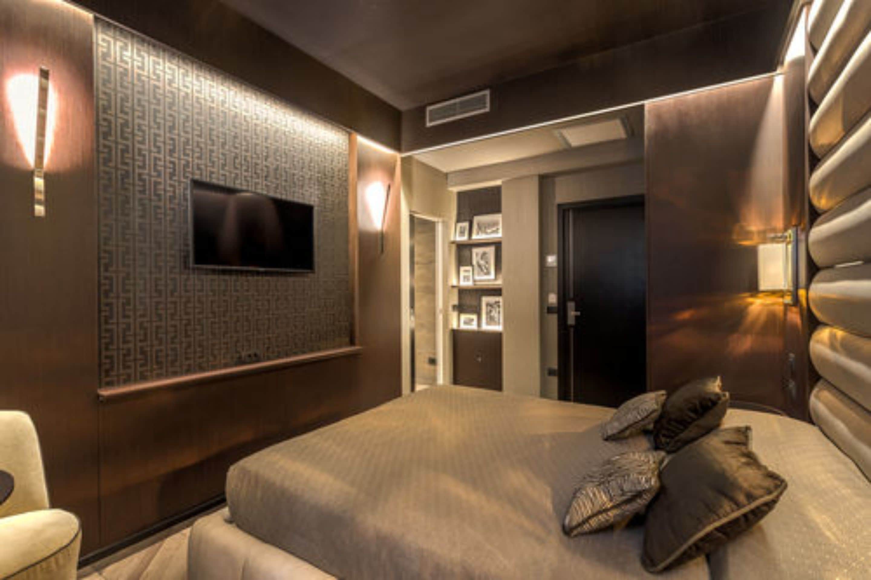 A modern bedroom with a large bed, a flat-screen TV mounted on the wall, and a door leading to another room.