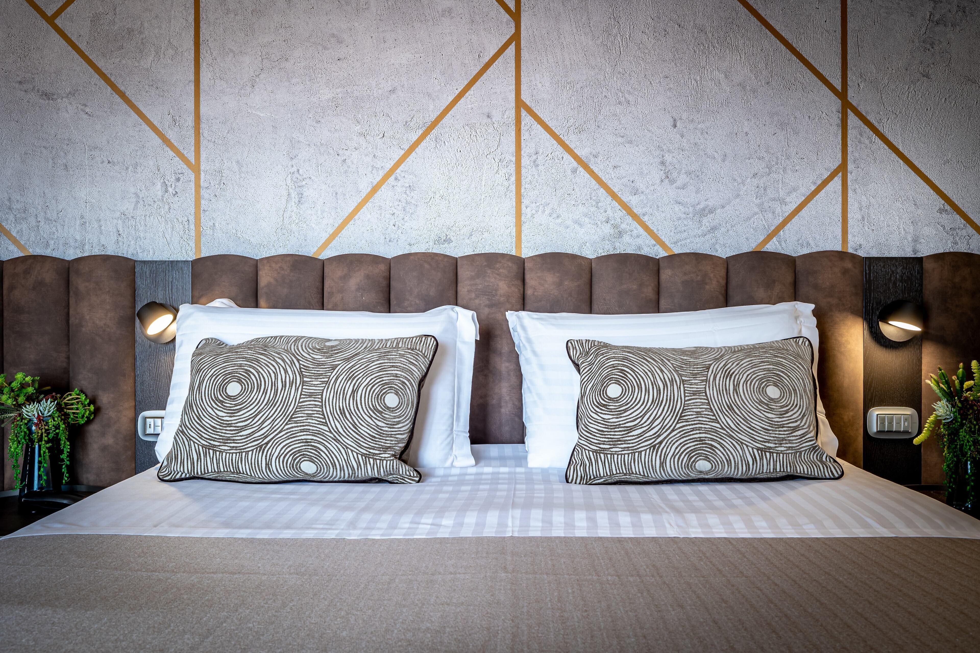 Une chambre d’hôtel avec un grand lit, deux coussins à motifs, une tête de lit dotée d’un éclairage intégré et un mur texturé orné de motifs géométriques.