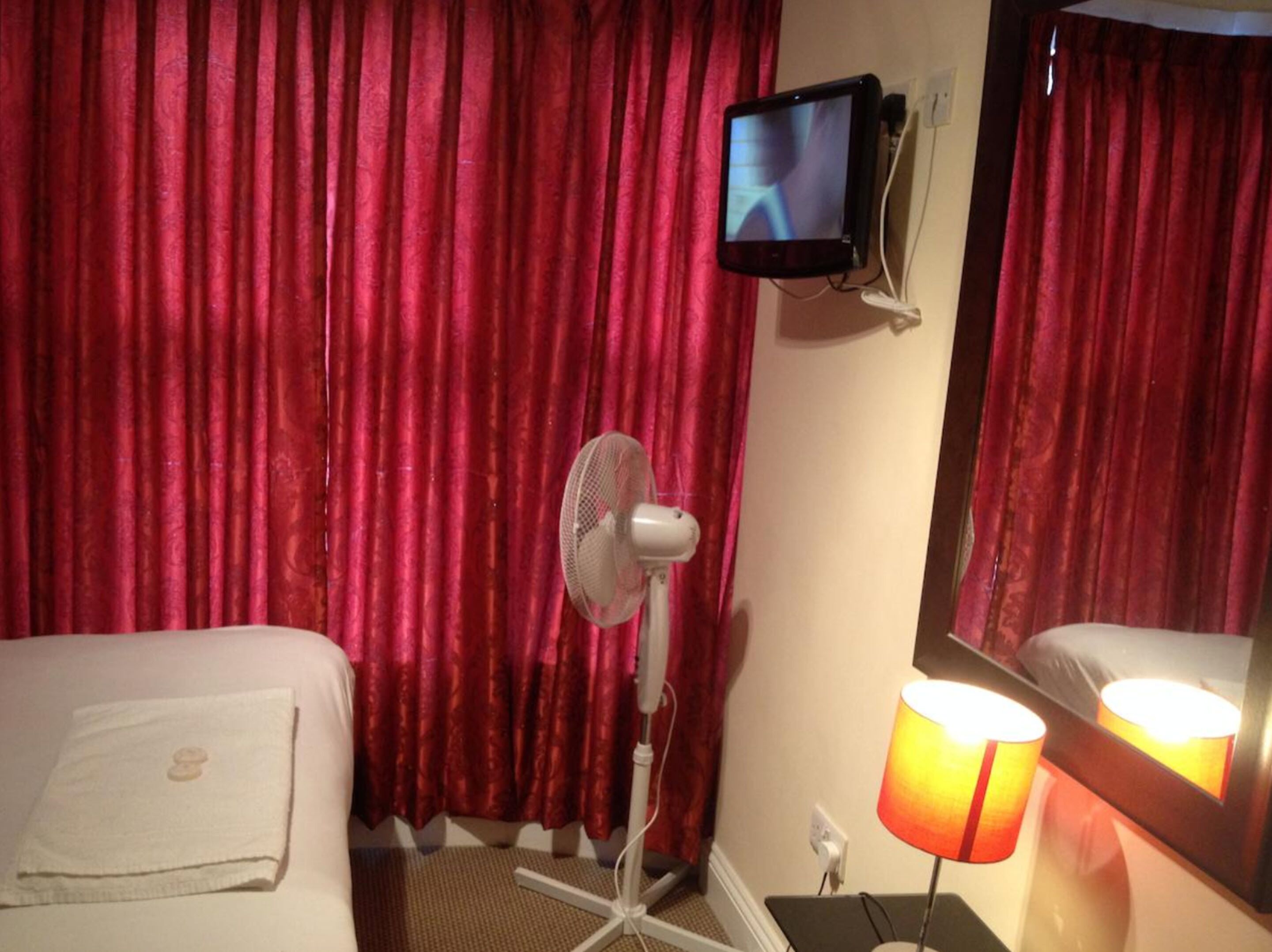 A hotel room with red curtains, a bed with a white towel, a standing fan, a television mounted on the wall, and a bedside lamp.