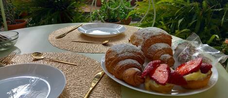 Une table dressée à l’extérieur, avec une assiette de croissants et de fraises, un verre transparent et un bol.