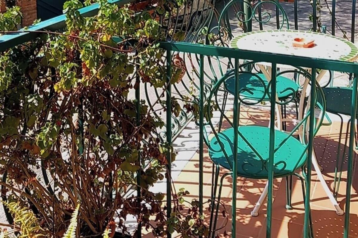 An outdoor seating area with green metal chairs and tables, surrounded by potted plants and hanging flower baskets.