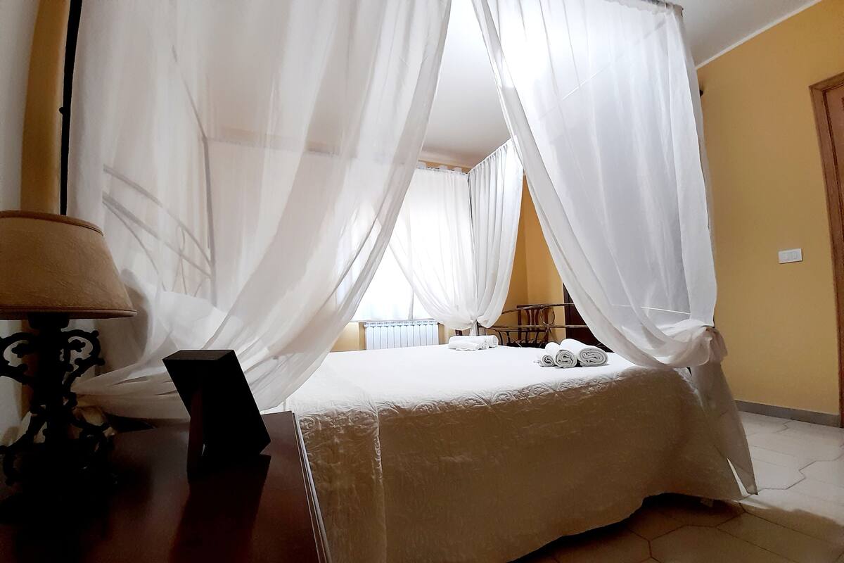 A bedroom with a four-poster bed, white bedding, and a bedside table with a lamp.