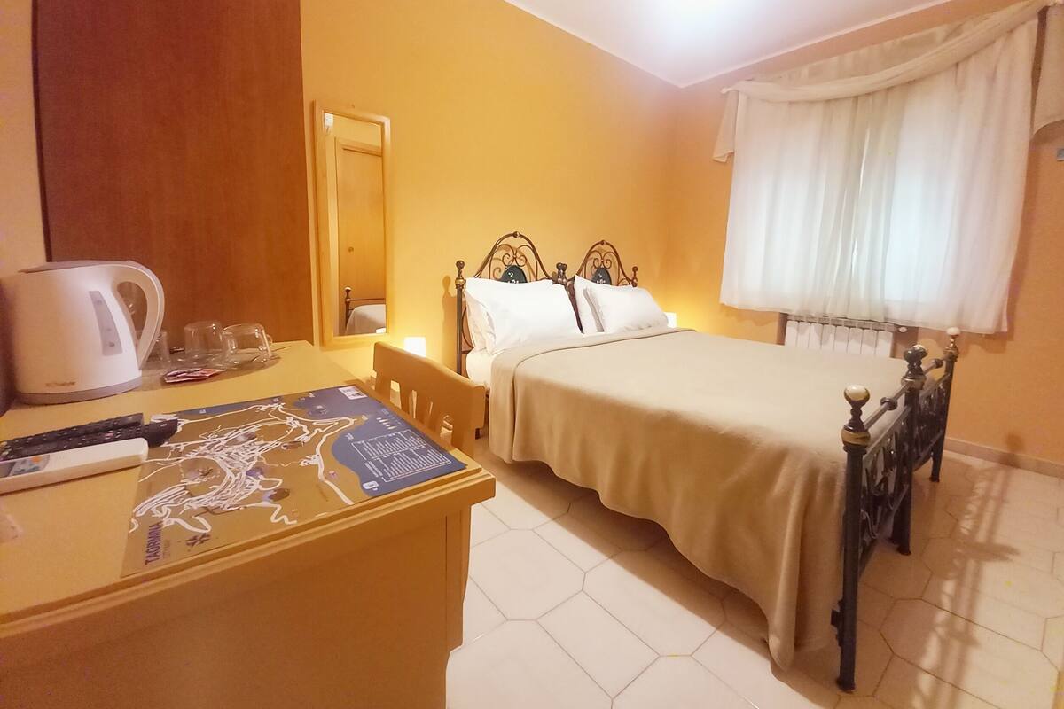 A hotel room with a bed, a desk with a kettle, a chair, and a window with curtains.