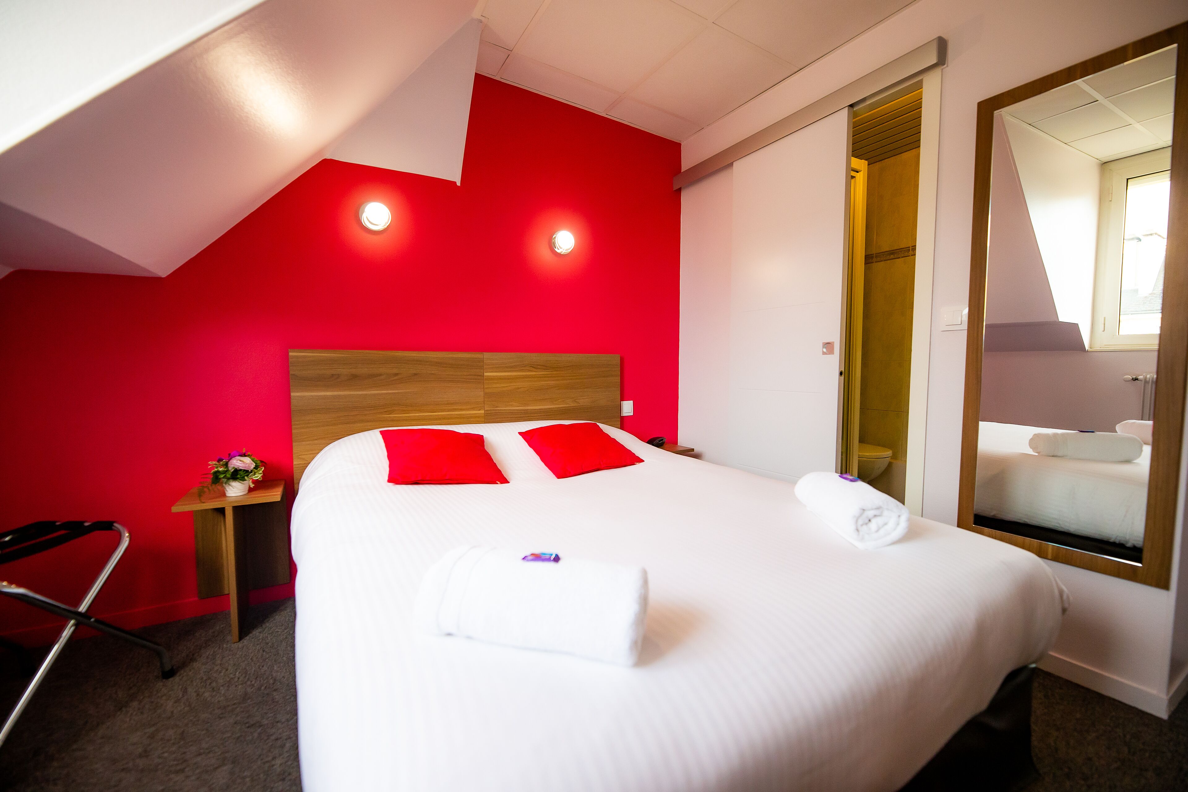 Une chambre d’hôtel avec un mur d’accent rouge, une tête de lit en bois, un lit blanc avec des oreillers rouges, une petite table de chevet agrémentée d’une fleur et un miroir avec un cadre en bois.