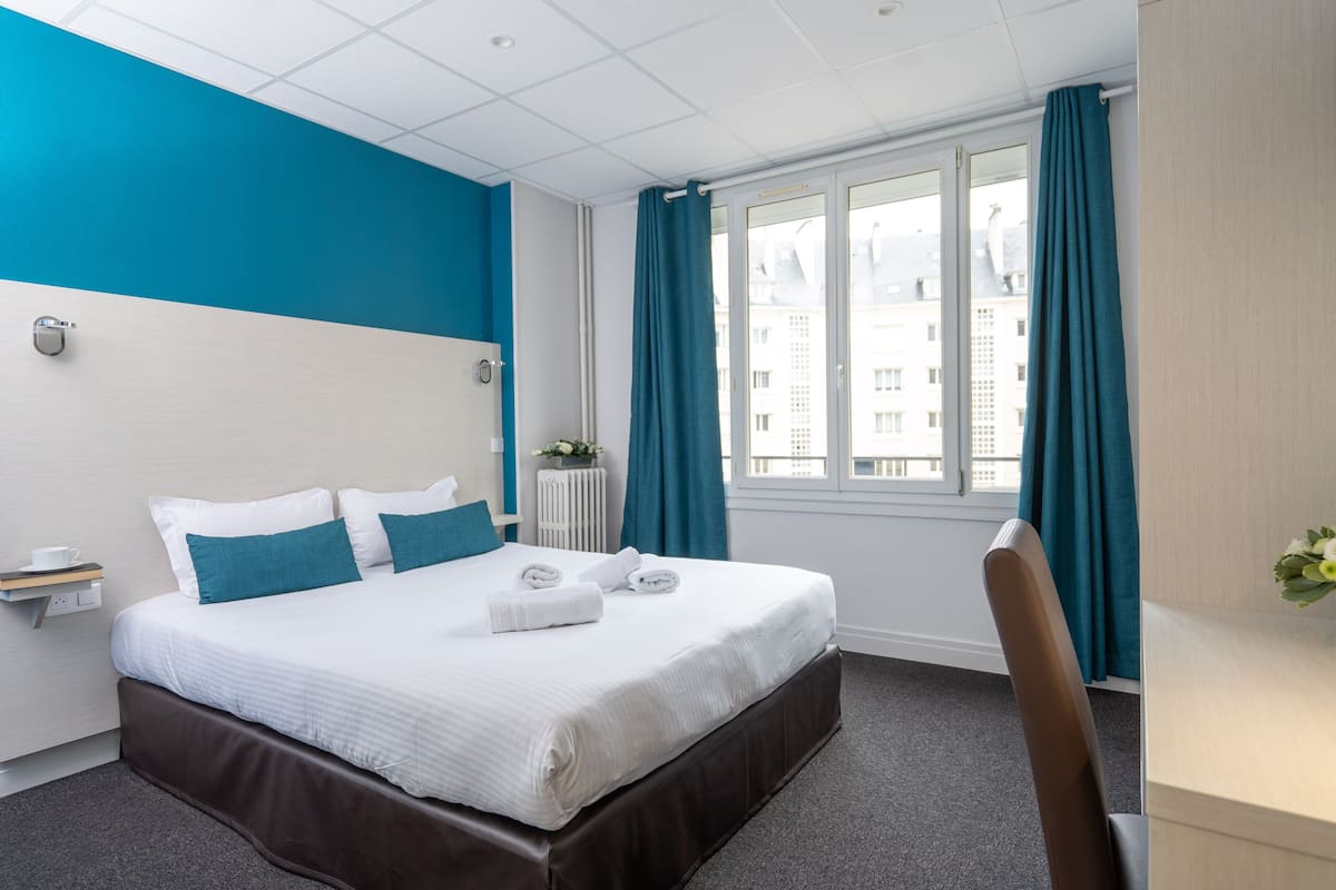 Habitación de hotel con una cama grande, un escritorio, una silla, una ventana con cortinas y vistas a edificios.