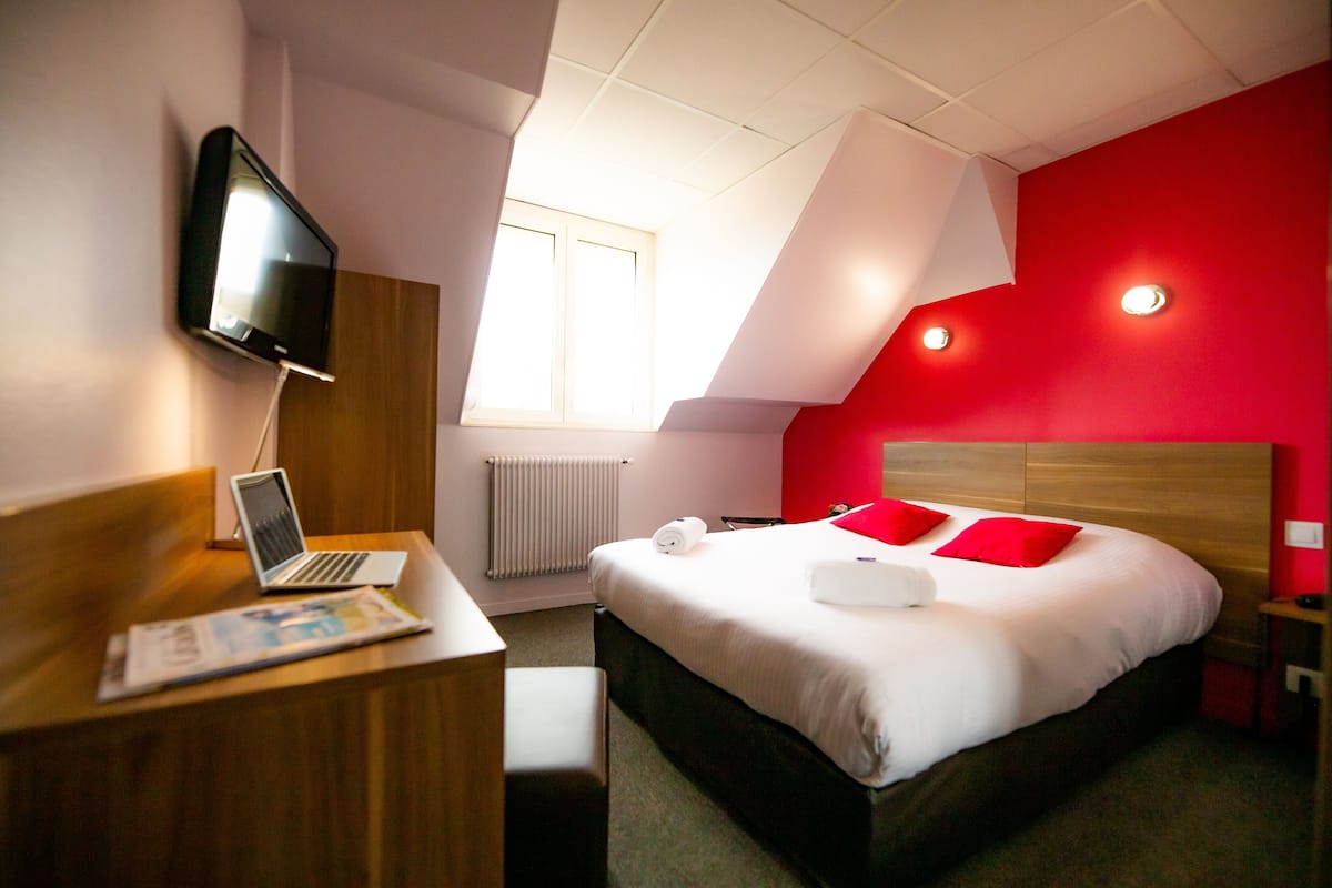 Una habitación de hotel moderna con una cama, un escritorio con una computadora portátil, un televisor de pantalla plana y una pared de acento roja.