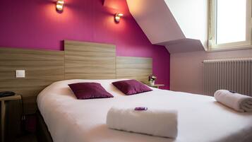Una habitación de hotel moderna con una cama grande, dos almohadas moradas, un edredón blanco y una toalla doblada con cuidado.