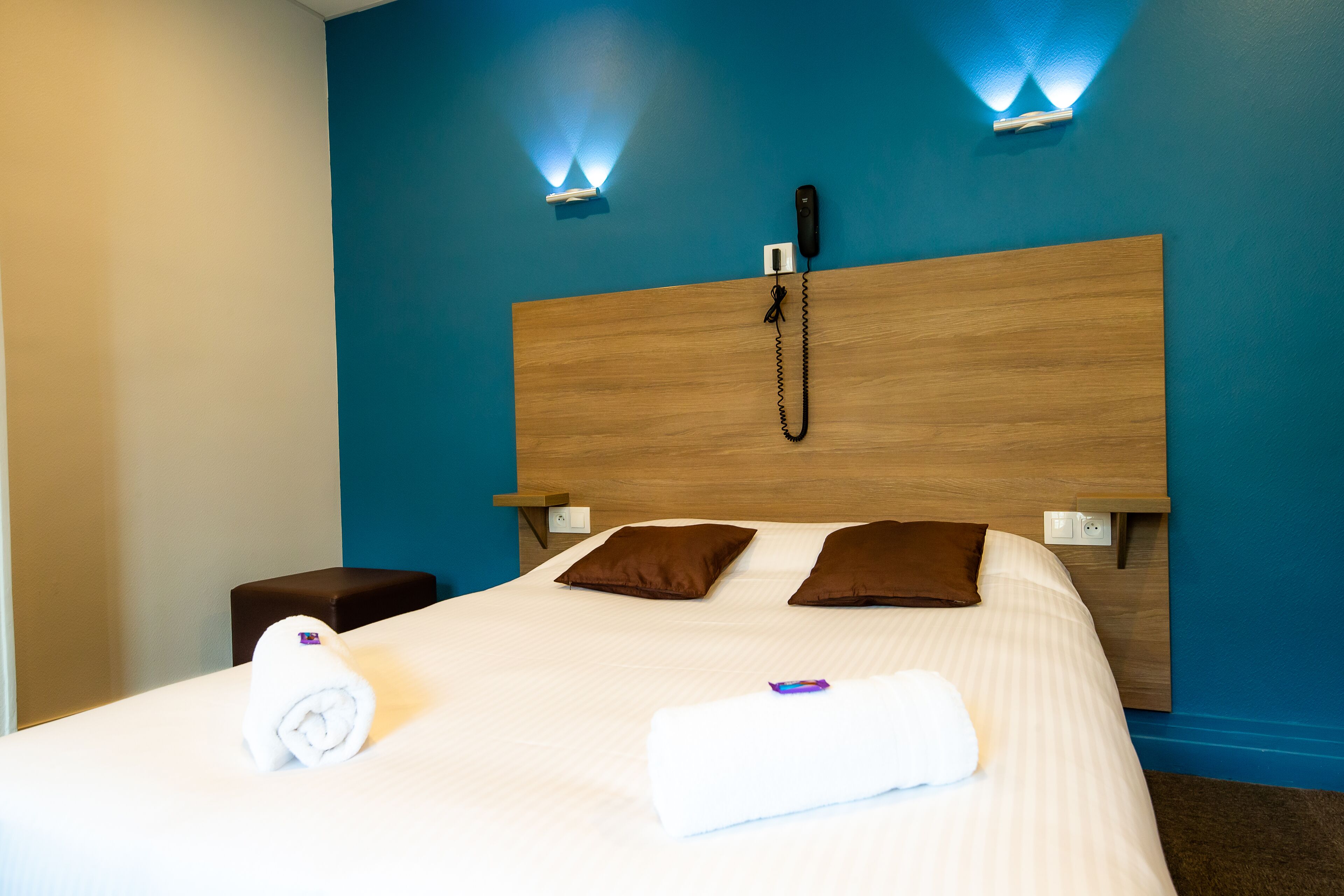 Habitación de hotel con cabecera de madera, dos almohadas marrones, una colcha blanca, una toalla blanca enrollada y un teléfono en la mesita de noche.
