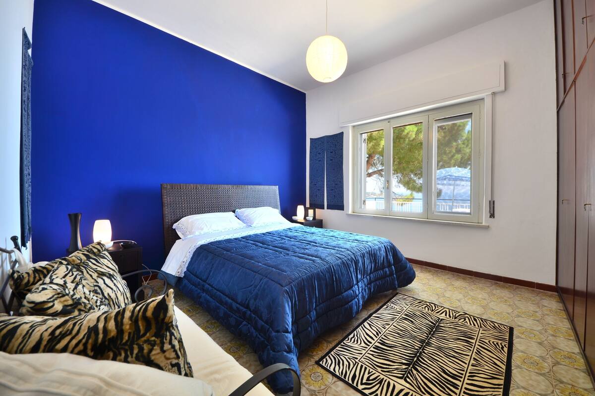 Une chambre avec un mur bleu, un lit orné d’un coussin à motifs zébrés, une table de chevet avec une lampe et une fenêtre donnant sur des arbres.
