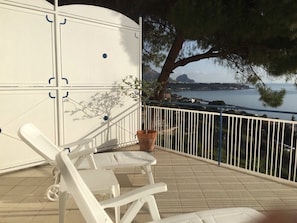 Un balcon avec des chaises blanches et un pot de fleurs, d’où l’on peut admirer un plan d’eau et un paysage lointain.