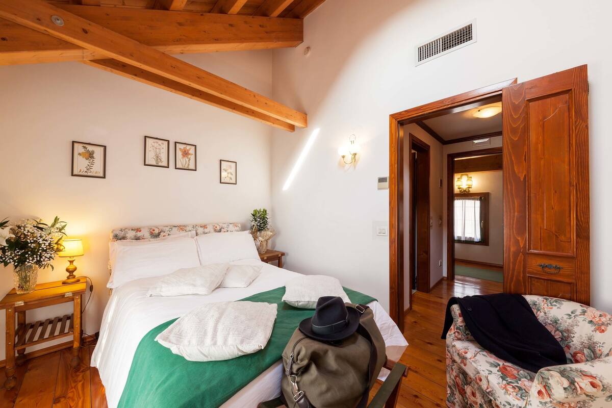 A bedroom with a bed, a nightstand, a lamp, a floral chair, and a wooden door.
