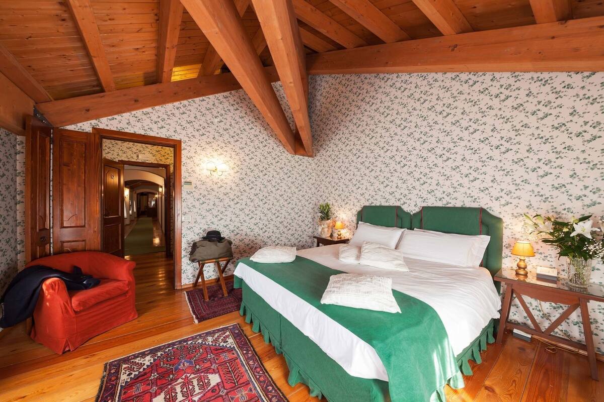 A room with a wooden ceiling, a bed with green bedding, a red chair, a nightstand with a lamp, and a floral wallpaper.