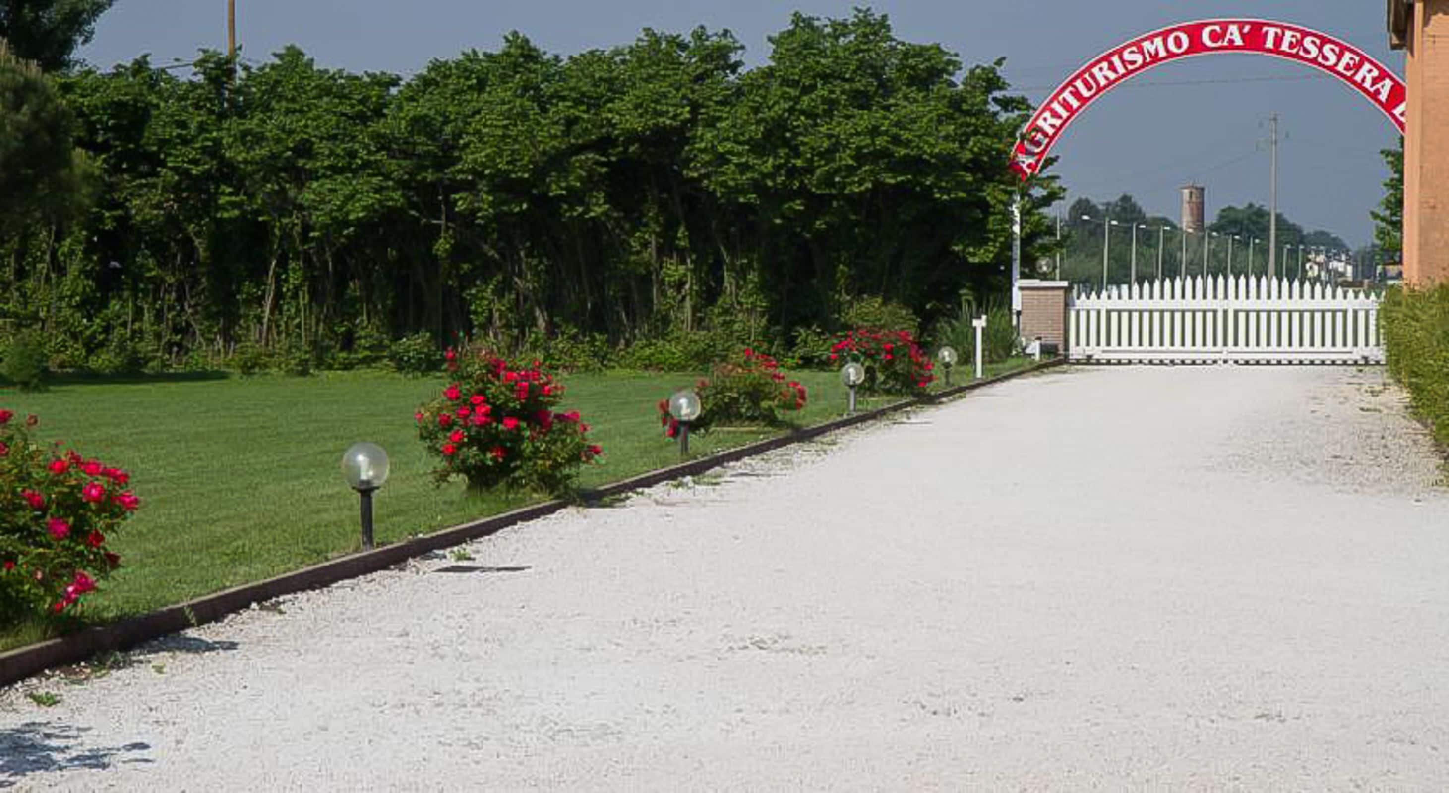 A gravel driveway leading to a white fence with a red banner reading "AGRITURISMO CA' TESSERA."