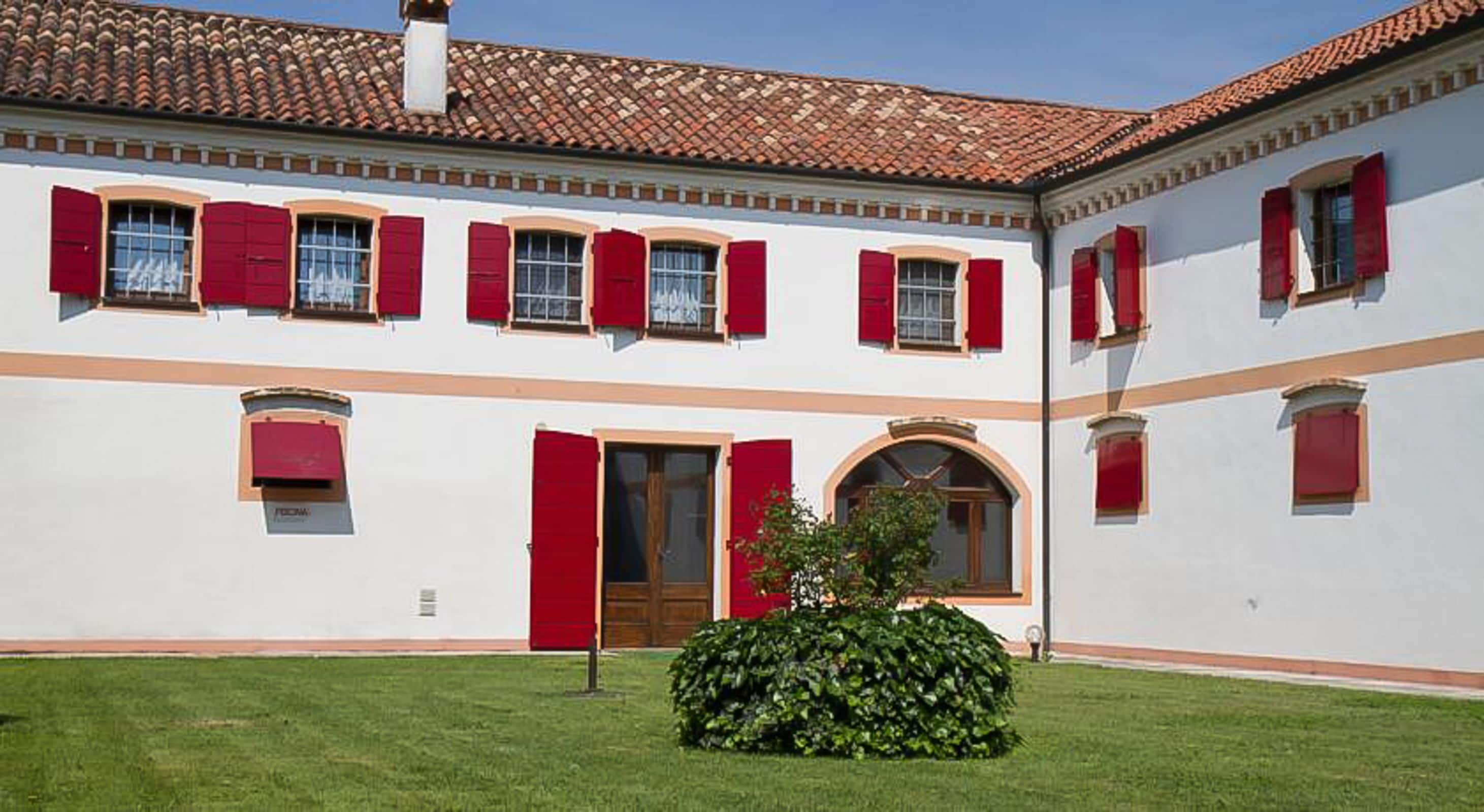 A white building with red shutters and a tiled roof. There is a small garden with a bush in the foreground.