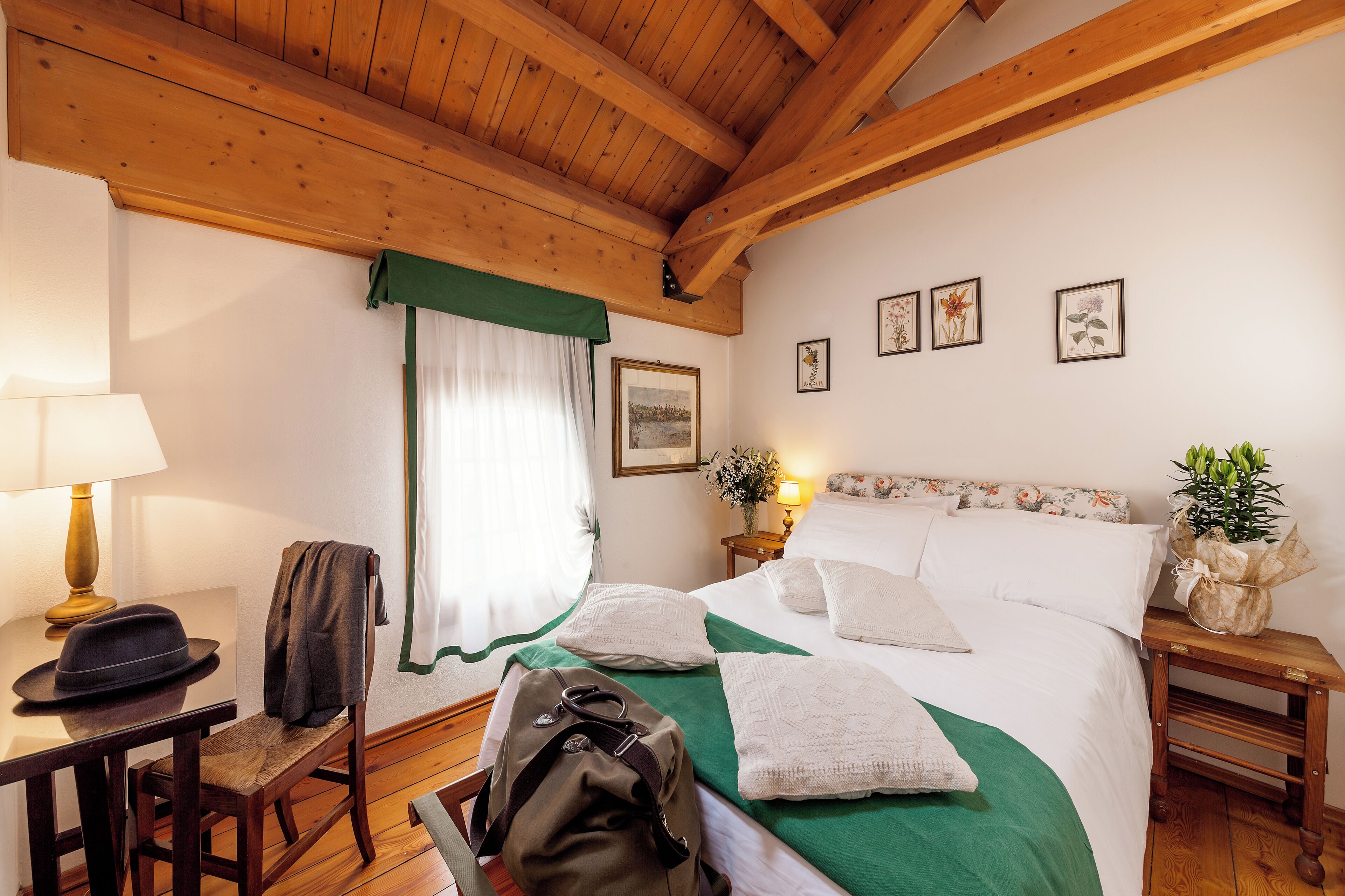 A hotel room with a wooden ceiling, a bed with white linens, a green bedspread, a nightstand with a lamp, a chair with a hat, and a backpack on the floor.