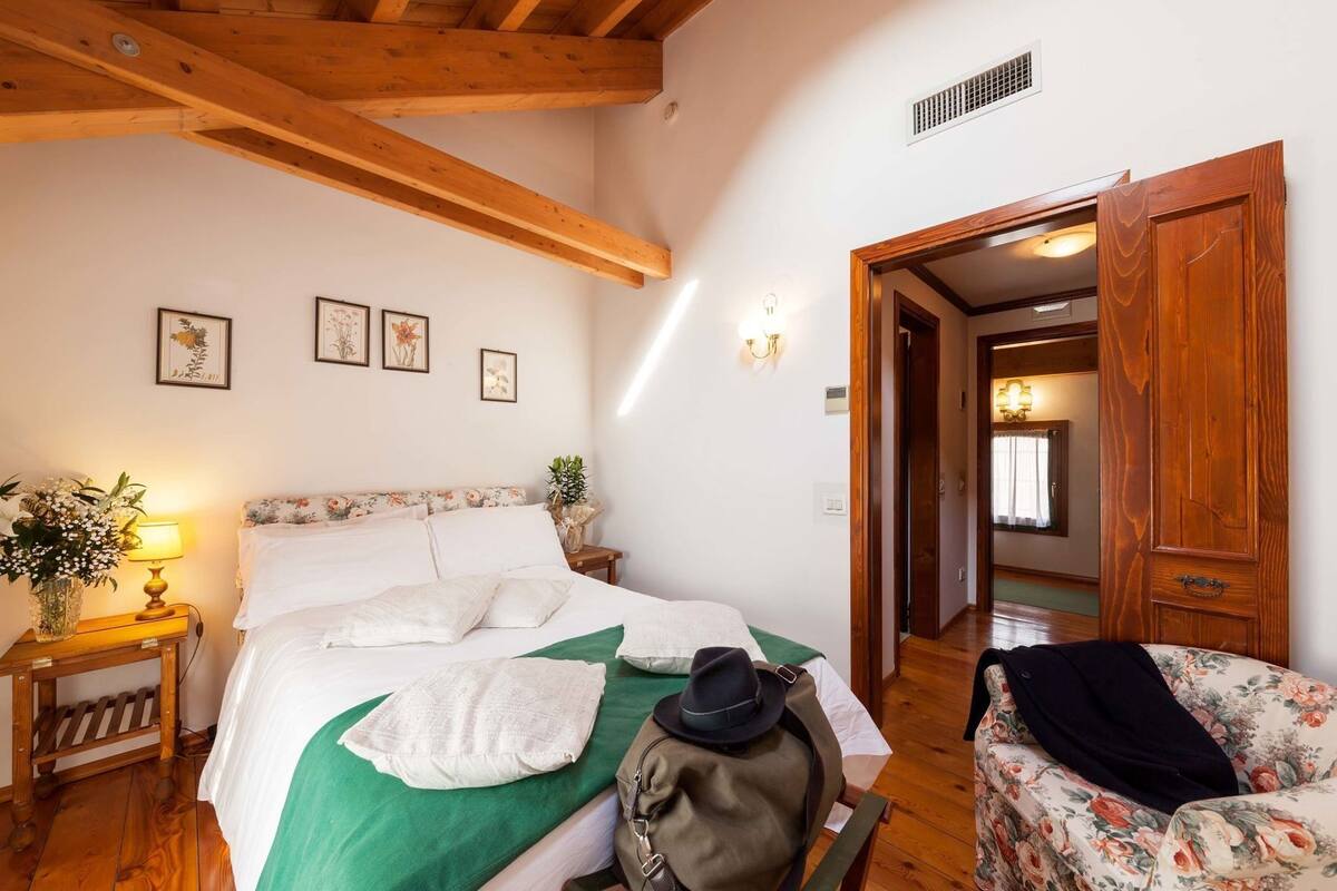 A bedroom with a wooden ceiling, a bed with white linens, a nightstand with a lamp, and a floral chair.