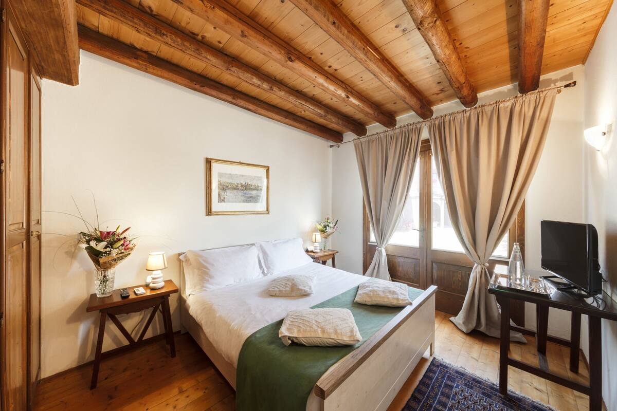 A bedroom with a wooden ceiling, a bed with white linens, a bedside table with a lamp and a vase of flowers, and a television on a desk.