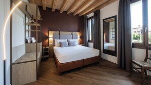 Un dormitorio con cama, suelo de madera, cabecero, mesita de noche, una silla y una ventana con cortinas.