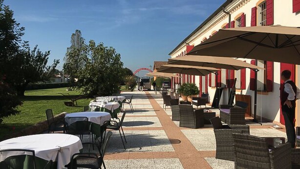 Un patio al aire libre con mesas y sillas, un edificio con contraventanas rojas y un cielo despejado.