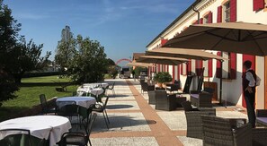 Un patio al aire libre con mesas y sillas, un edificio con contraventanas rojas y un cielo despejado.
