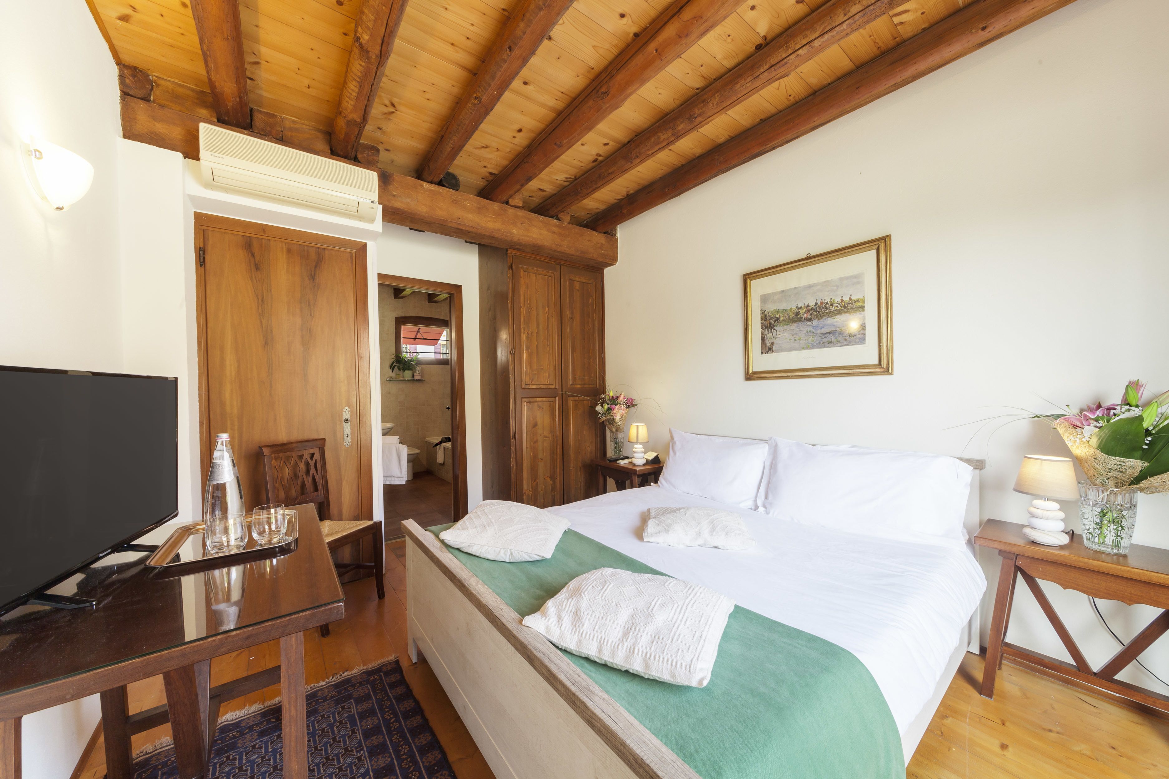 Habitación de hotel con techo de madera, cama con ropa blanca, televisor sobre una mesa de madera y una mesita auxiliar con lámpara y flores.