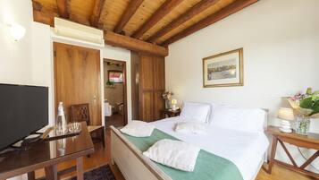 Habitación de hotel con techo de madera, cama con ropa blanca, televisor sobre una mesa de madera y una mesita auxiliar con lámpara y flores.