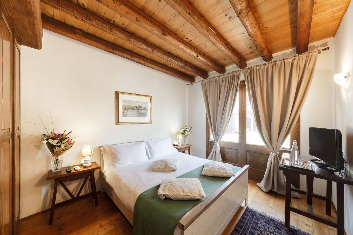 A bedroom with a wooden ceiling, a bed with white linens, a bedside table with a lamp and flowers, and a desk with a computer.