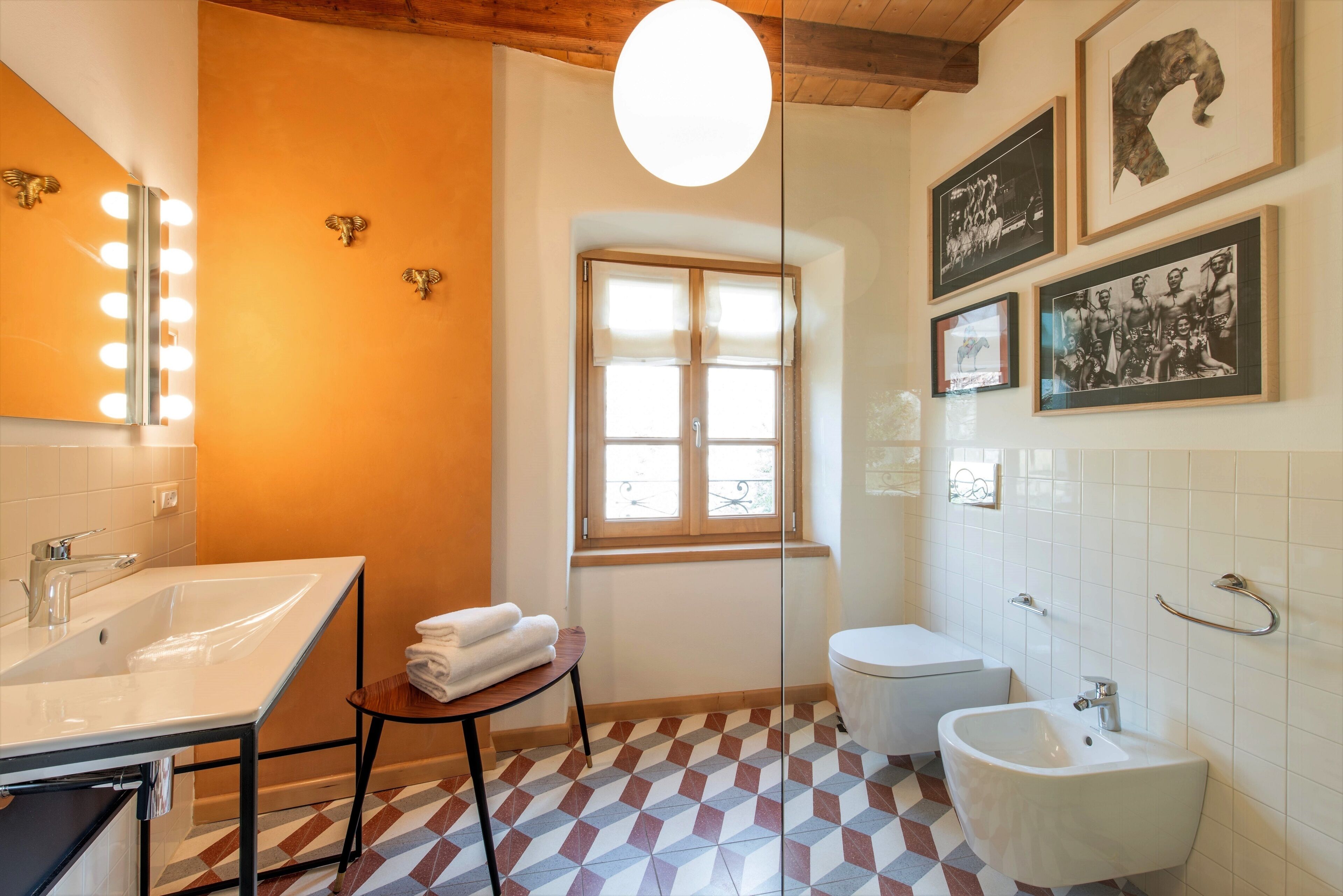 A bathroom with a large mirror, vanity, sink, and bidet. There are towels on a stool, a window, and framed pictures on the wall.