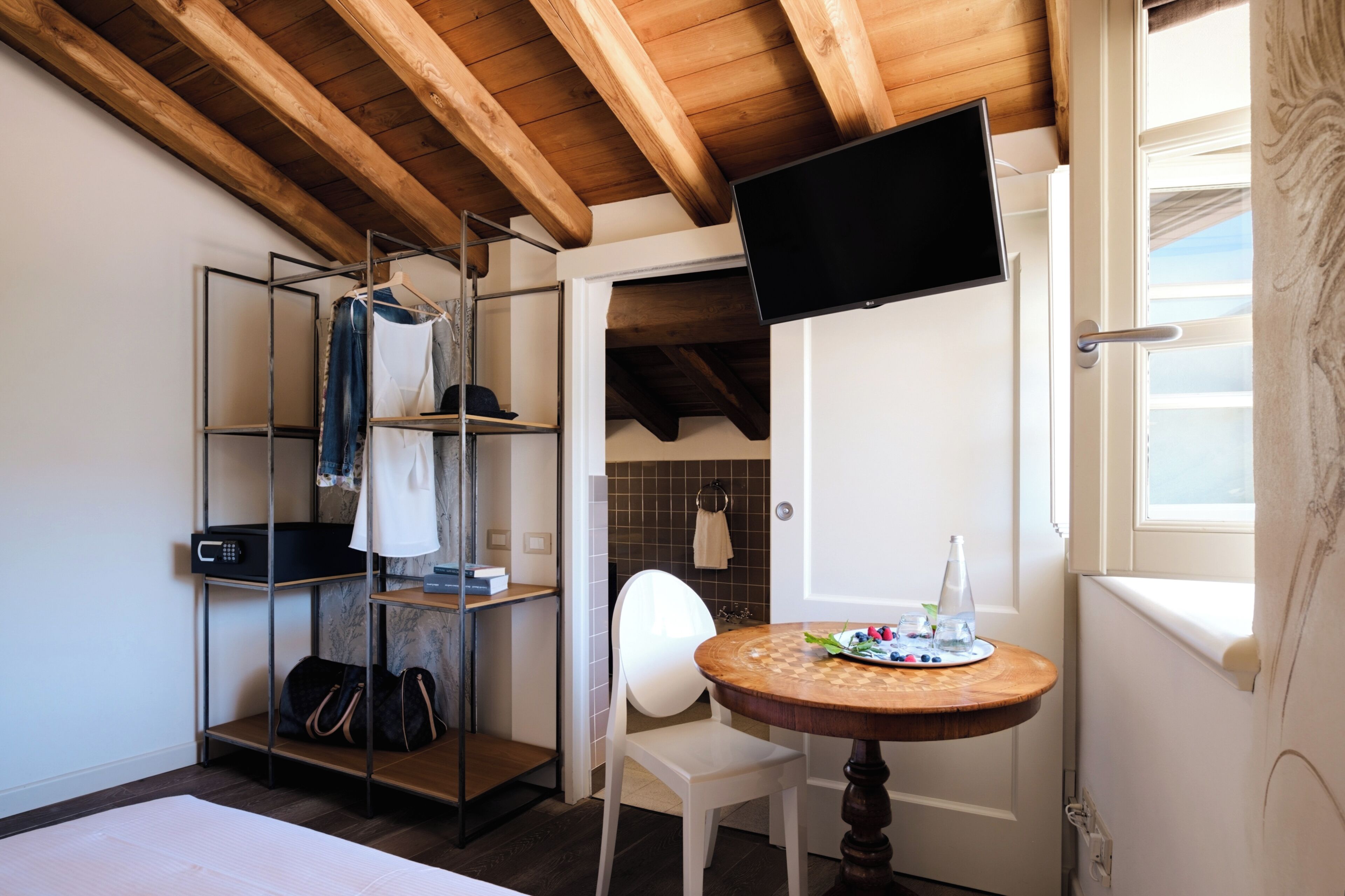 Una camera compatta con soffitto in legno, una TV montata a parete, un piccolo tavolo rotondo con un piatto di frutta, una sedia bianca e un appendiabiti con abiti appesi.