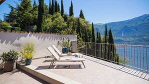 Une terrasse offrant une vue sur un lac et des montagnes, avec une table et des chaises, des plantes en pot et une rambarde.