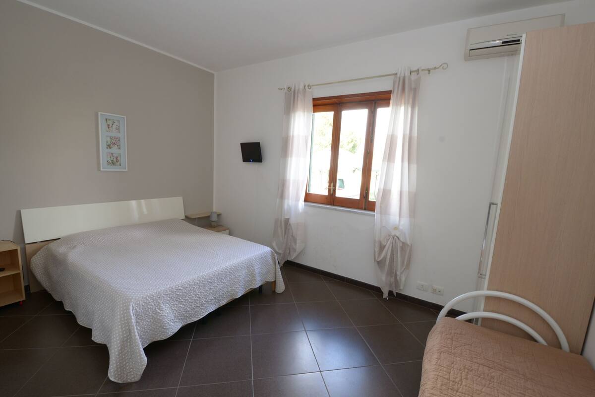 Une chambre à coucher comprenant un lit, une table de chevet, une chaise, une fenêtre avec des rideaux et un appareil de climatisation.