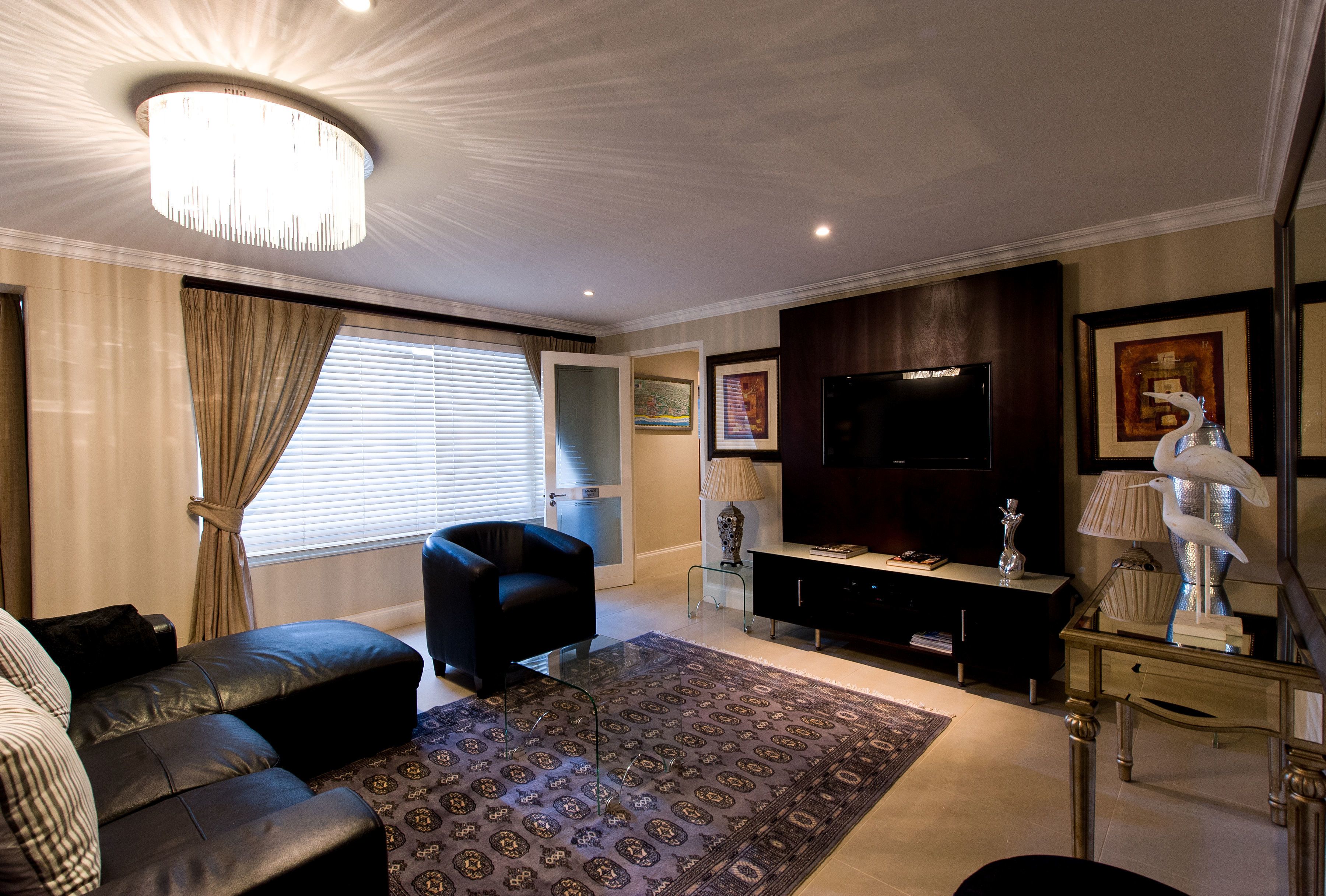 Un salon moderne avec un canapé en cuir noir, un tapis à motifs, une table basse en verre, un téléviseur à écran plat et une sculpture d’oiseau décorative.