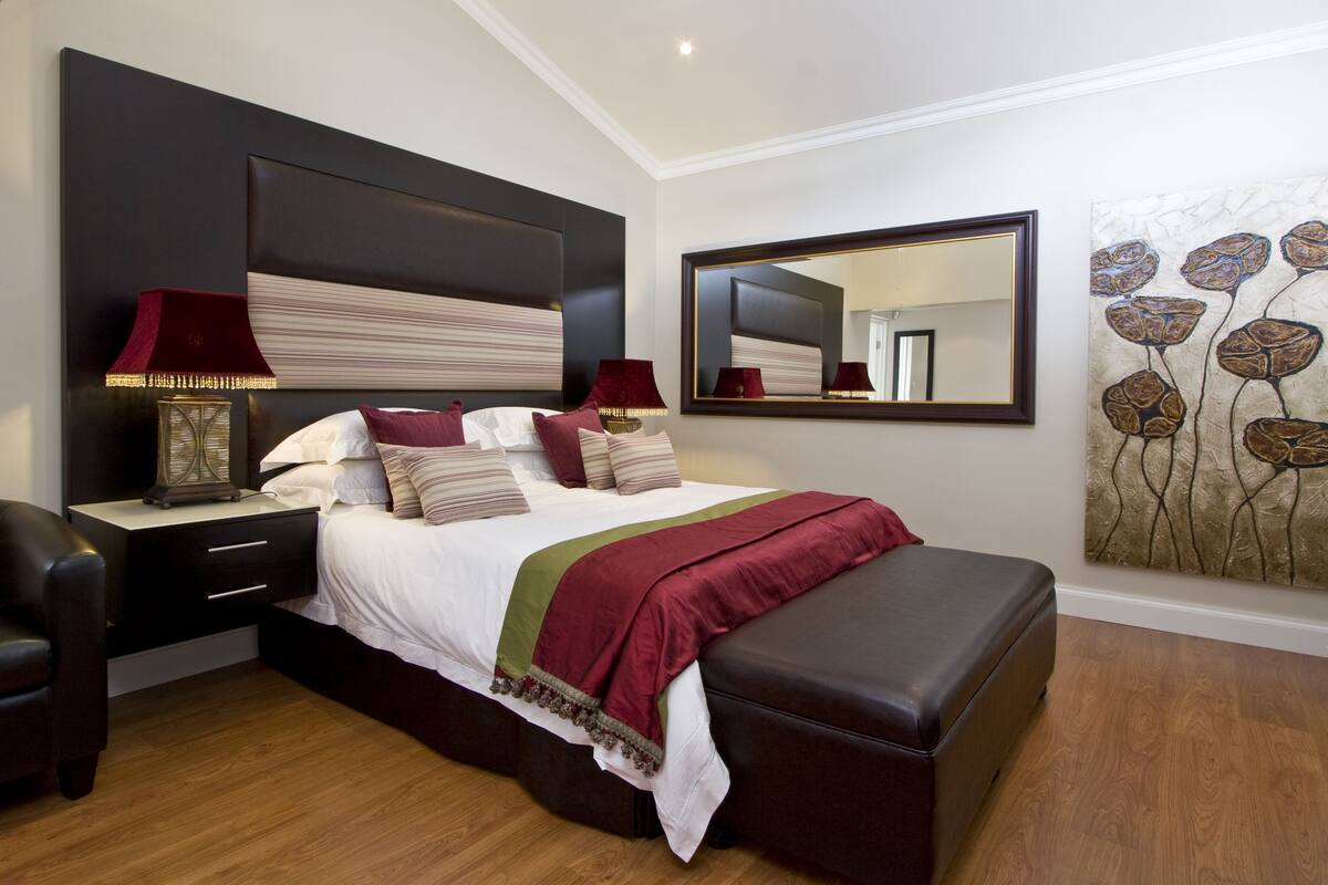 A bedroom with a large bed, bedside tables, a nightstand, a chair, a bench, a mirror, and a wall art piece.