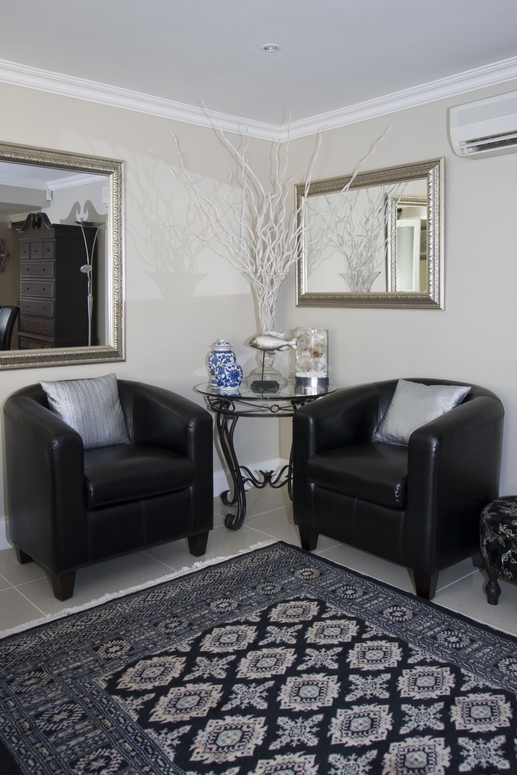 A room with two black leather armchairs, a patterned rug, a glass coffee table with decorative items, and two large mirrors on the walls.