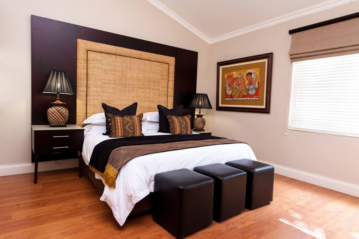 A bedroom with a large bed, two bedside tables, a headboard, a framed artwork, and a window with blinds.