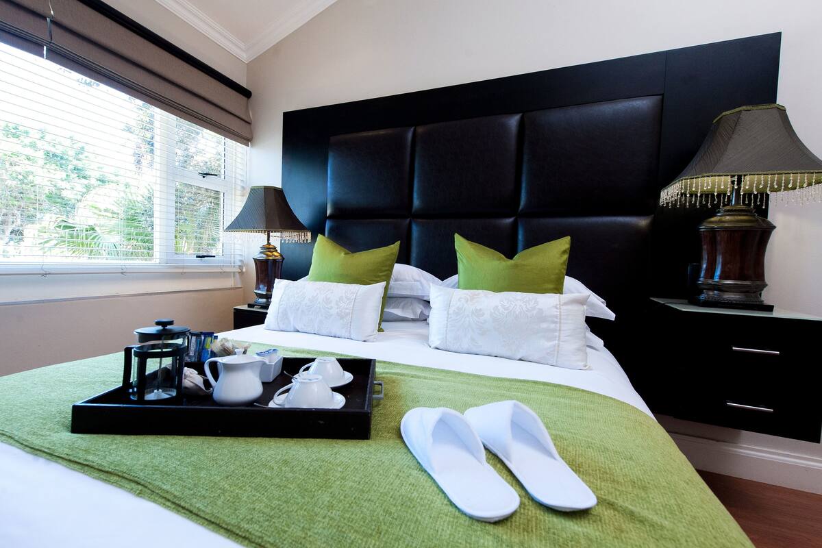 A neatly made bed with a black headboard, green and white pillows, a tray with a coffee maker, and slippers on a green bedspread.