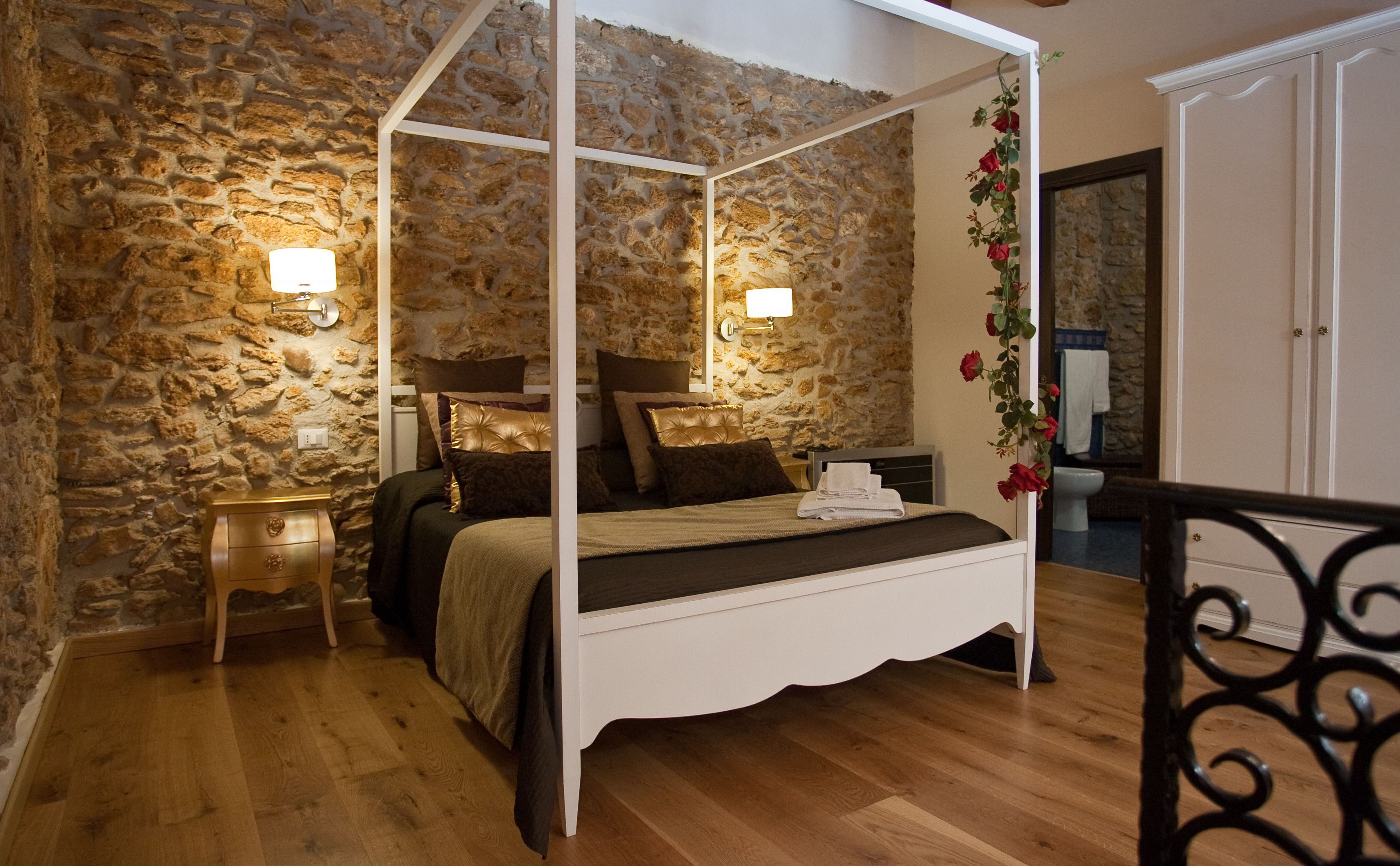 Un lit à baldaquin avec un mur en pierre en arrière-plan, une table de chevet en bois et une armoire blanche.