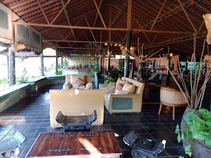 A spacious indoor area with a high ceiling, furnished with sofas and chairs, a wooden coffee table, and potted plants.